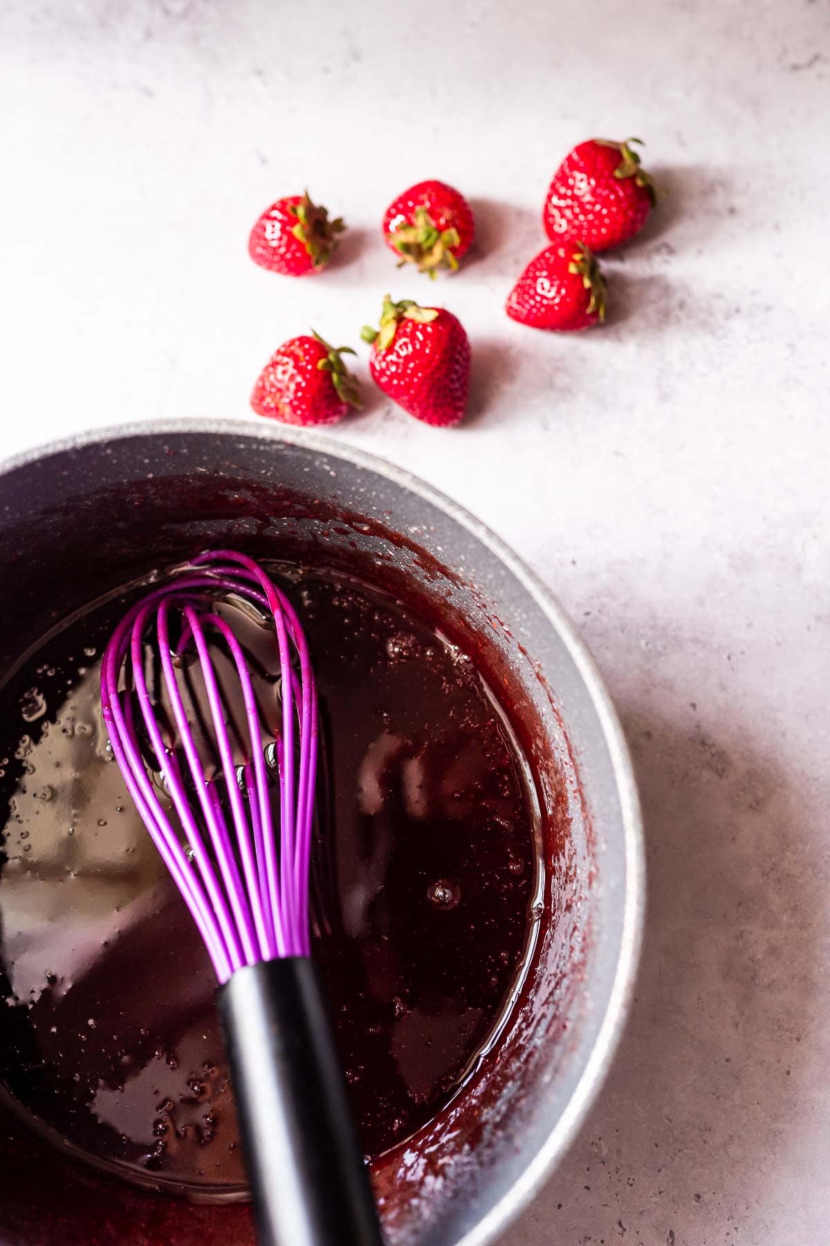 A purple whisk rests in a small saucepan filled with a red syrup.