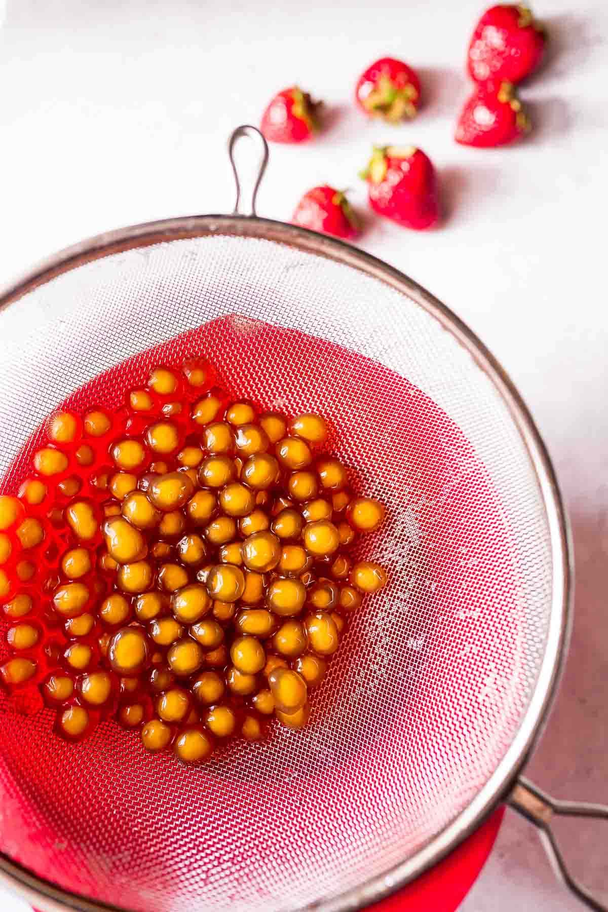 A mesh strainer filled with boba pearls rests on a bright red mixing bowl.