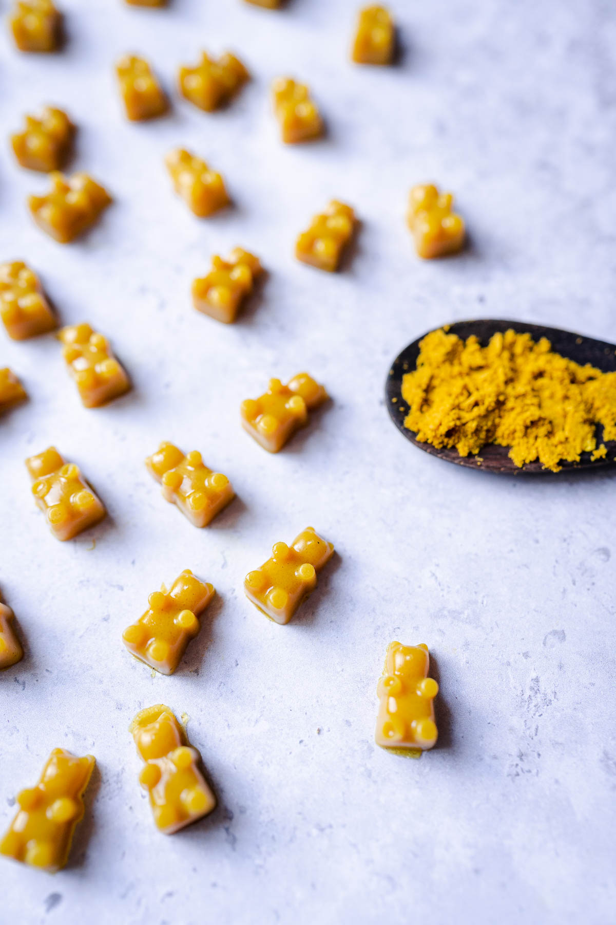 A small wooden spoon filled with golden milk powder rests next to a bunch of yellow gummy bears on a gray tabletop.