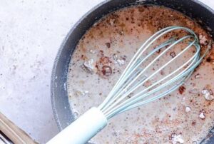 A whisk rests in a saucepan filled with a creamy vegan queso mixture of milk and spices on a light-colored countertop.