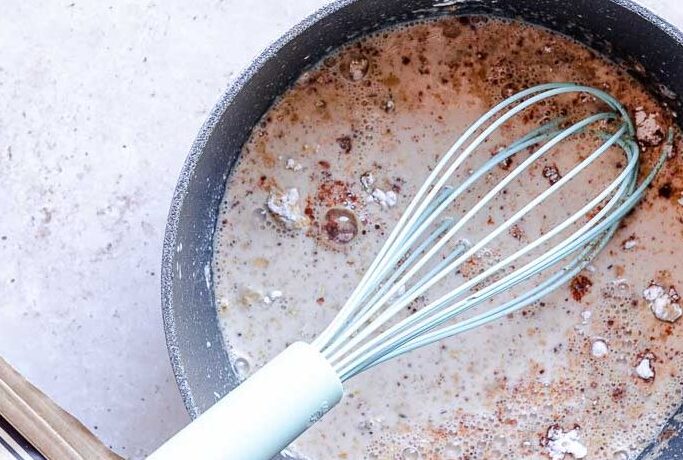 A whisk sticking out of a saucepan with a milk like substance.