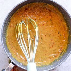 A whisk rests in a pan filled with thick, orange-brown vegan queso, dotted with green and red specks, on a light-colored surface.
