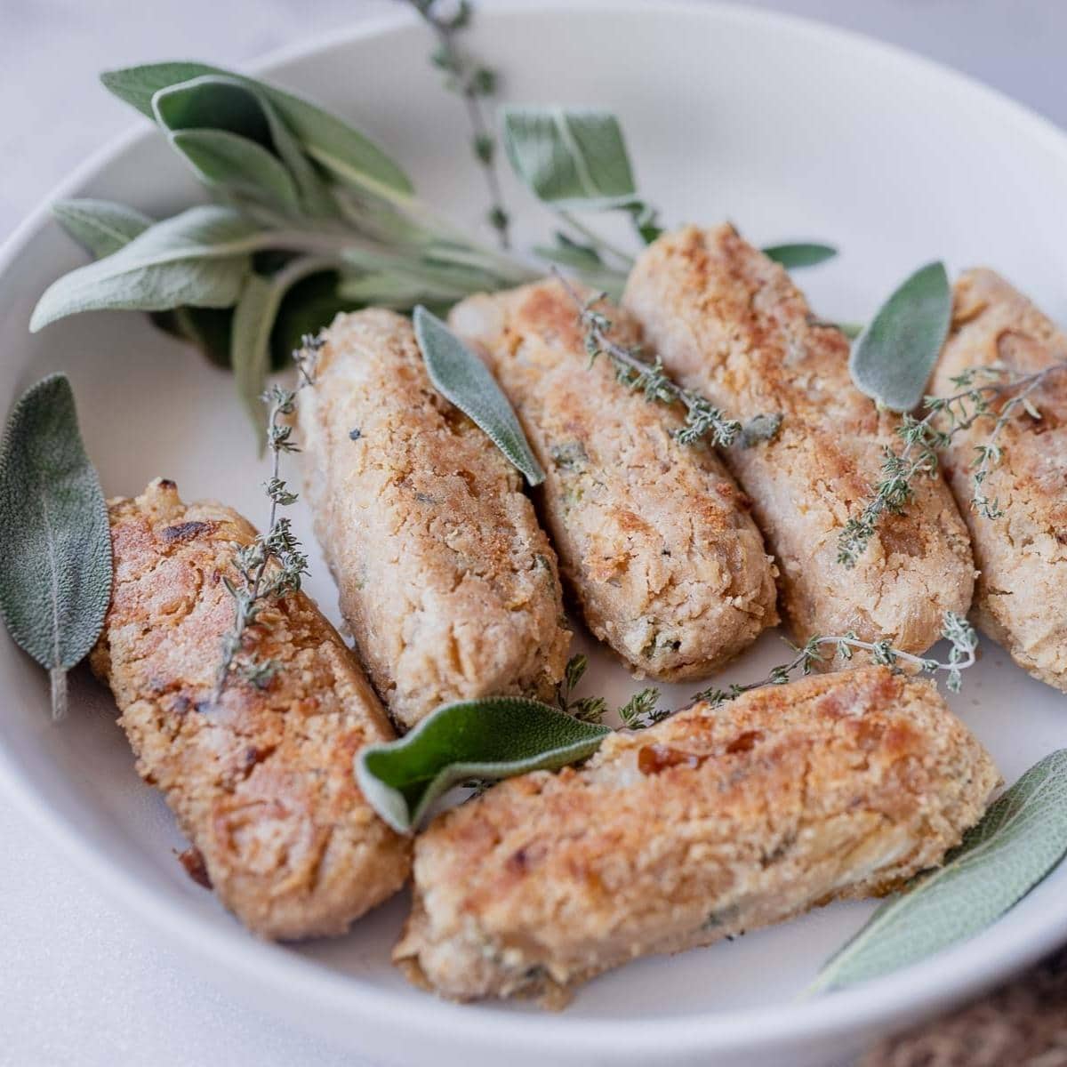 A white ceramic bowl filled with tan sausages and fresh herbs.