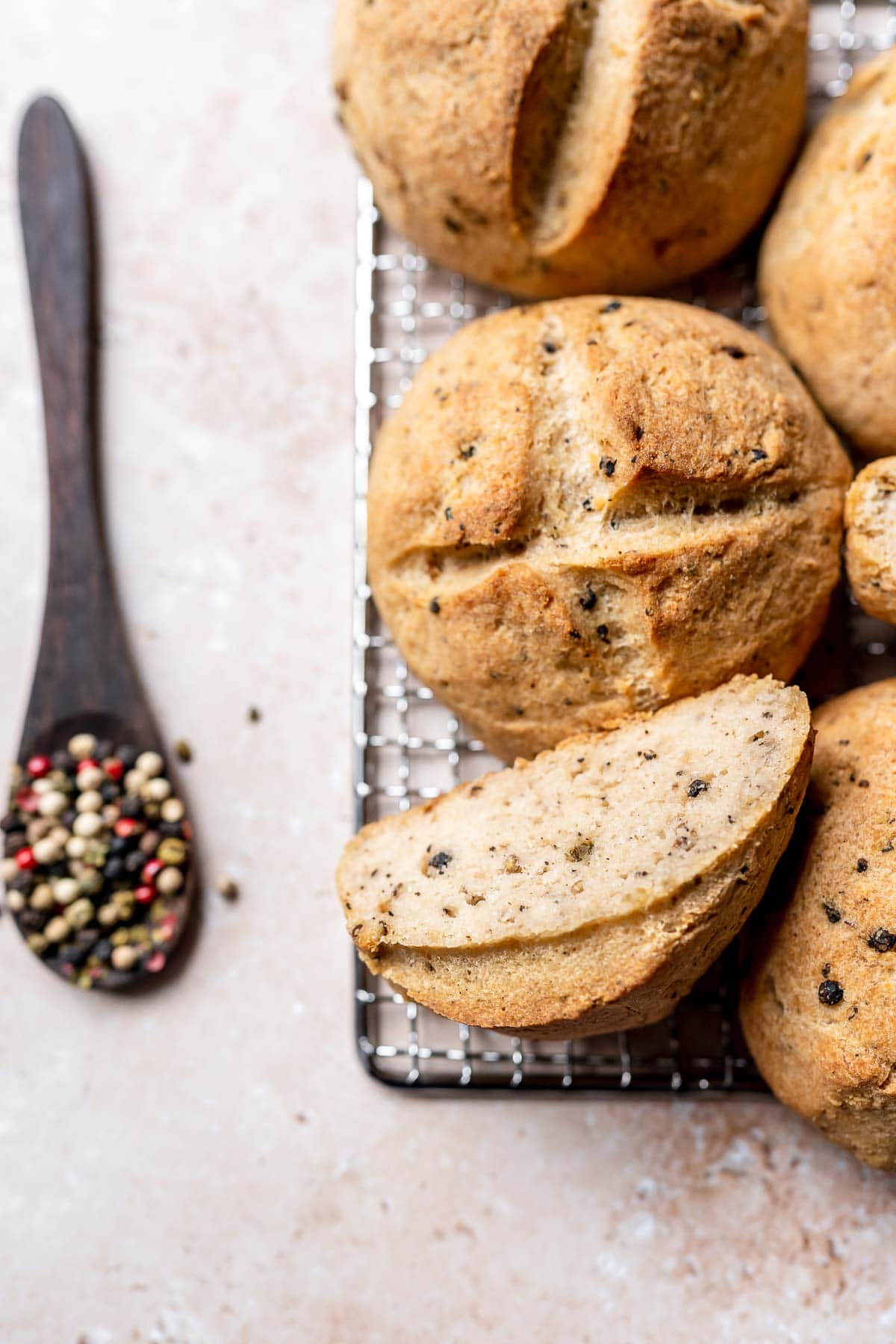 Freshly baked gluten free sourdough rolls resting on a cooling rack next to a wooden spoon filled with rainbow peppercorns.