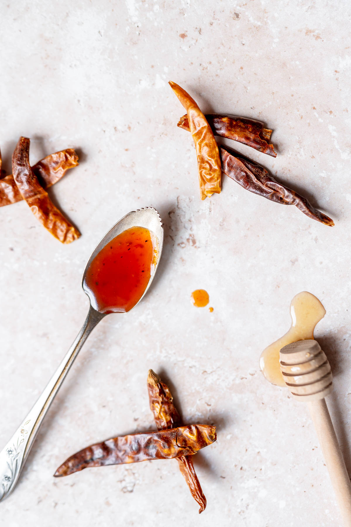 Top view of a silver spoon filled with a bright red sauce resting on a table surrounded by dried chiles and a wooden honey spoon dripping with honey.