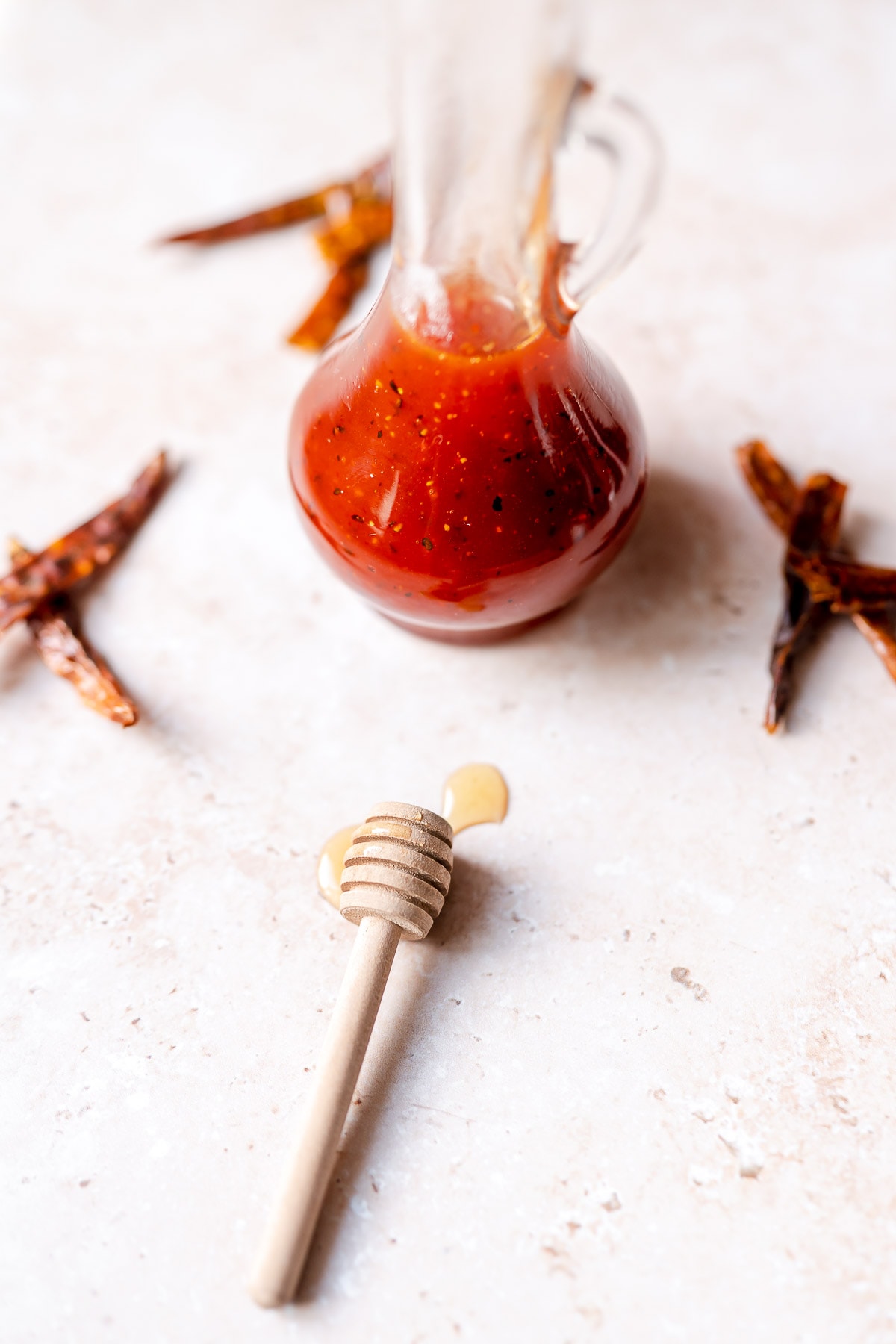 A wooden honey spoon dripping with honey rests next to a clear glass bottle filled with a bright vibrant red sauce.