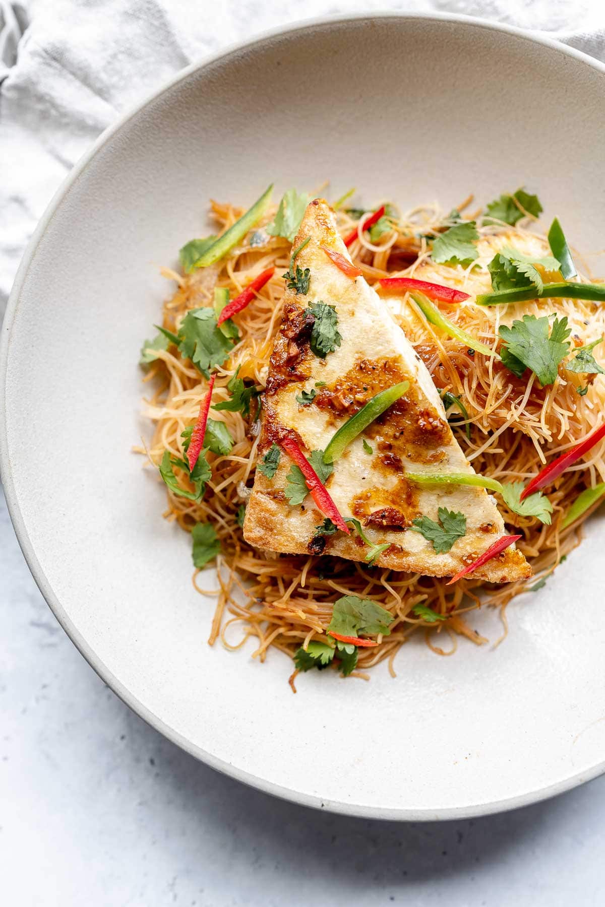 A triangular slab of tofu resting on a bed of thin noodles in a large tan bowl.