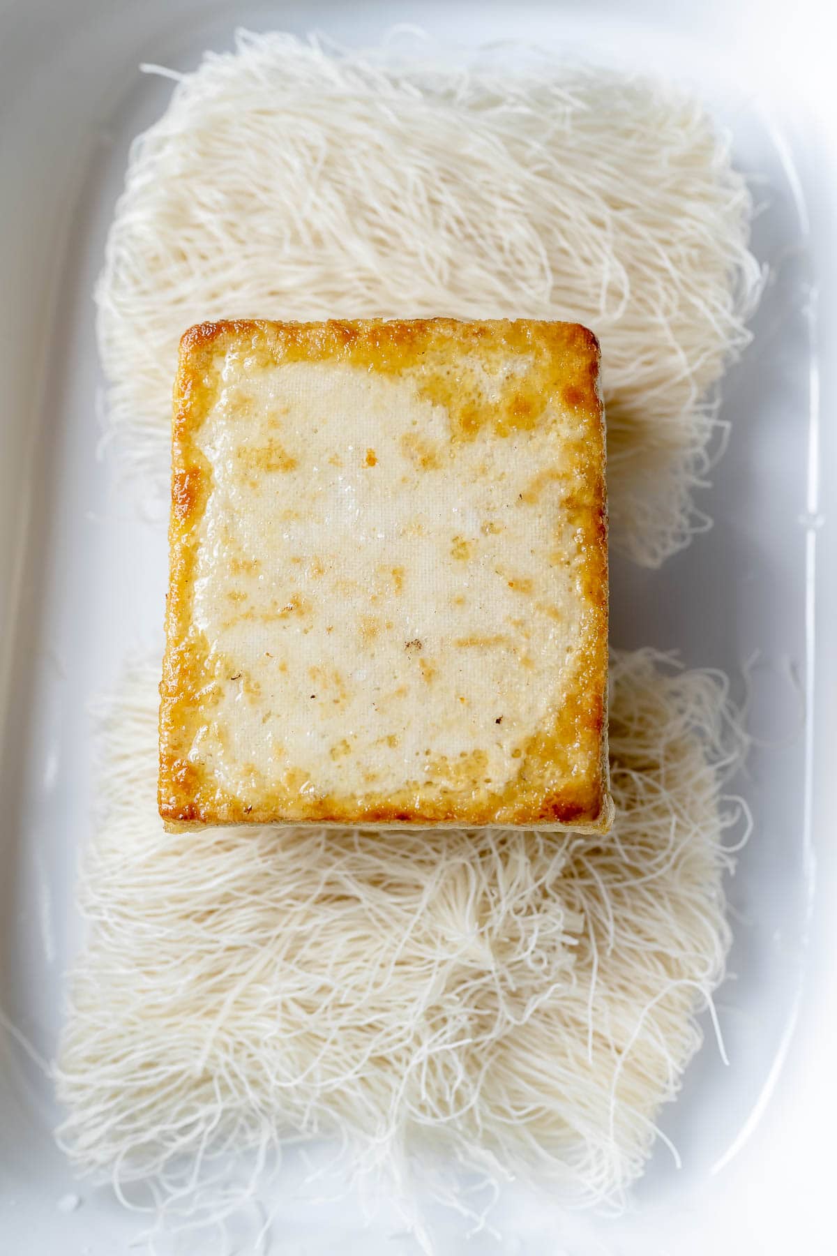 A golden block of tofu resting on dried noodle nests in a white baking dish.