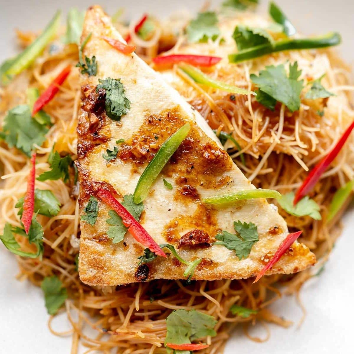 A triangle of white tofu topped with veggies resting on a bed of cooked brown noodles.