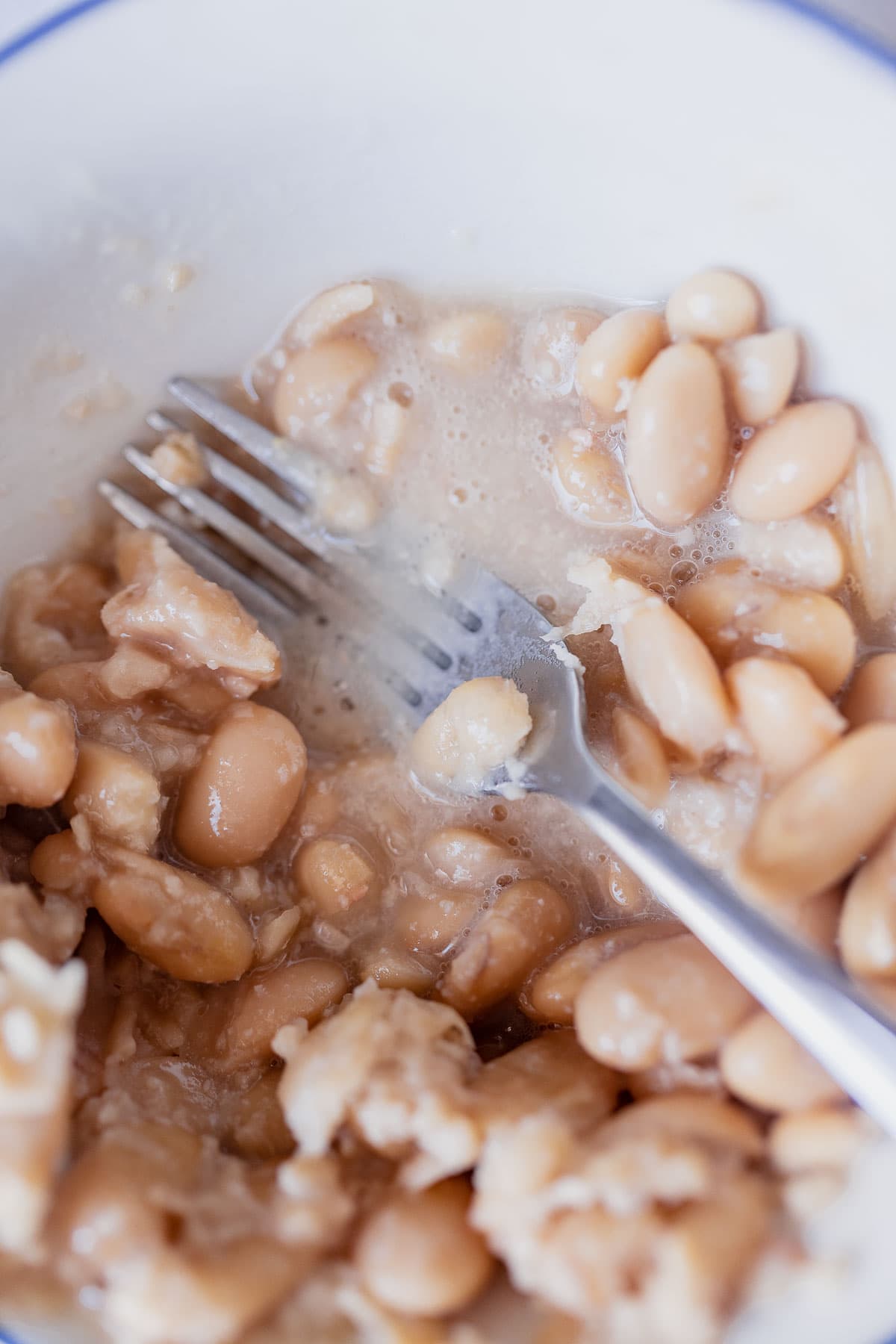 A silver fork mashing butter beans in a white bowl.