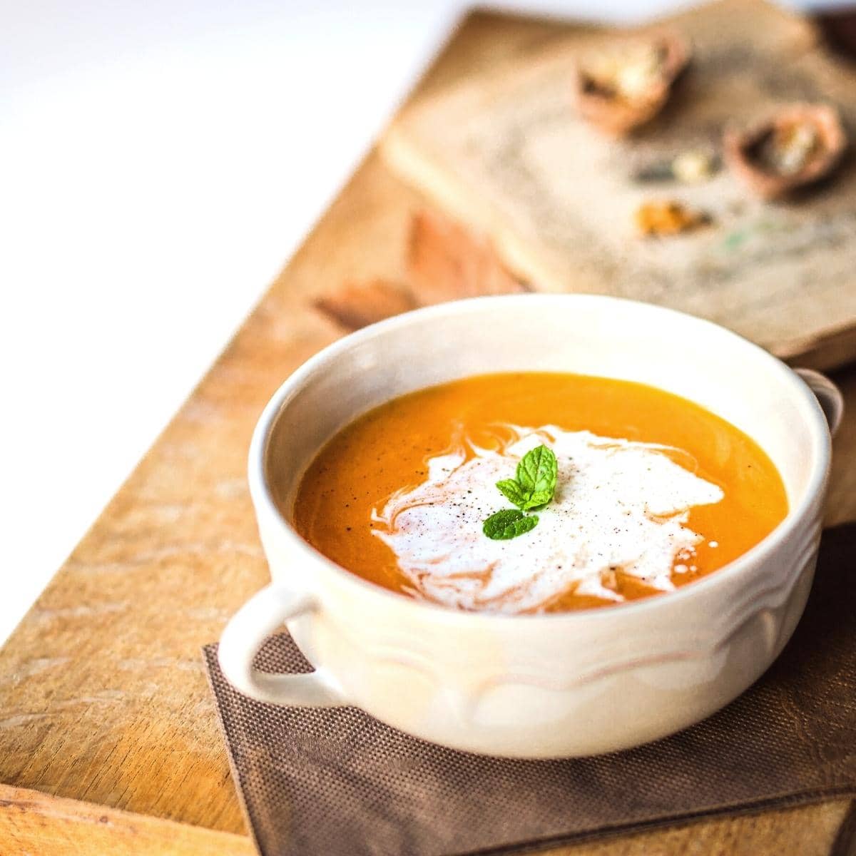 A white ceramic bowl filled with orange soup topped with white cream and fresh herbs.