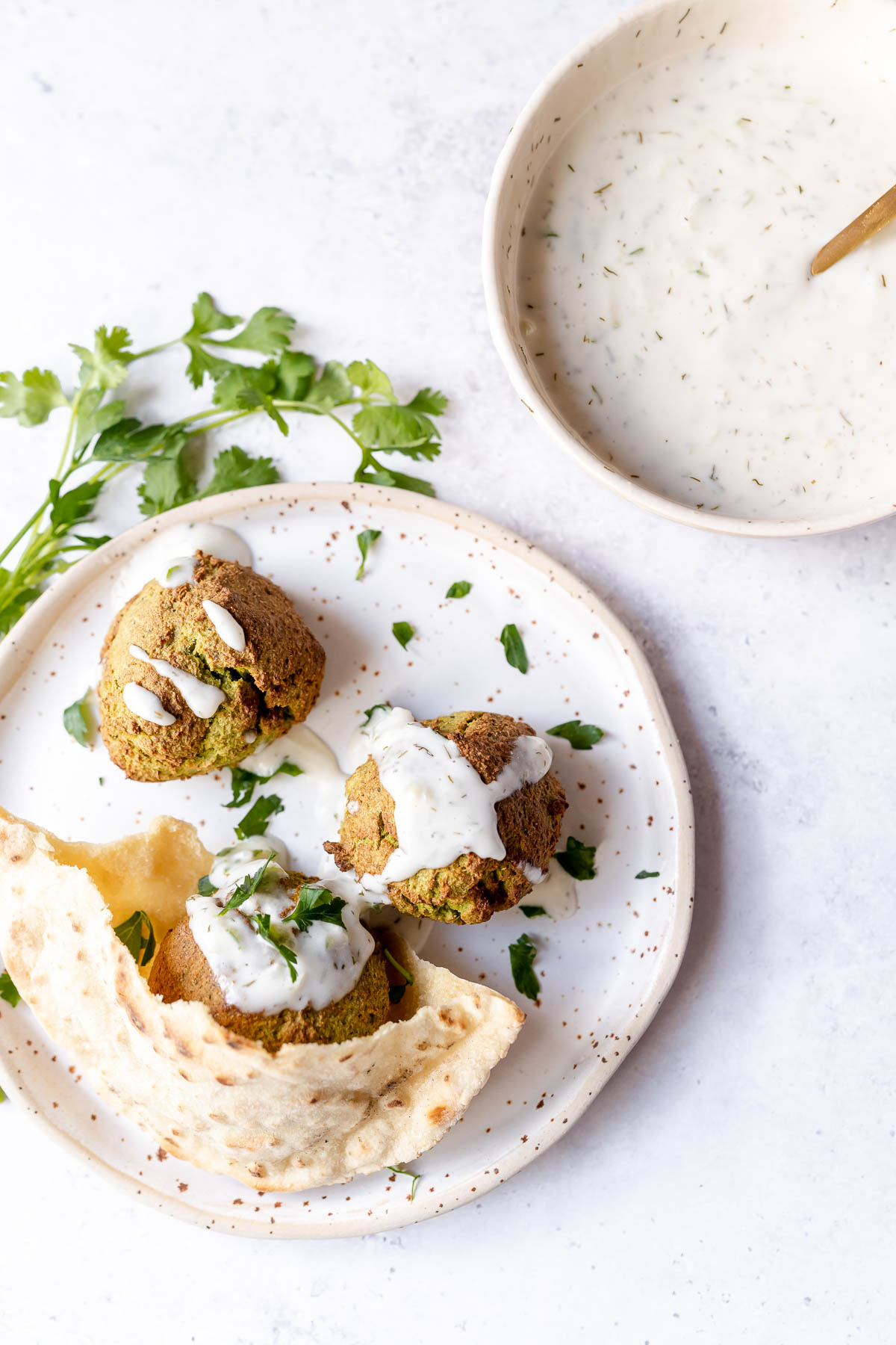 Top view of a white plate topped with golden falafel, fresh green herbs, white tzatziki sauce and pita bread.