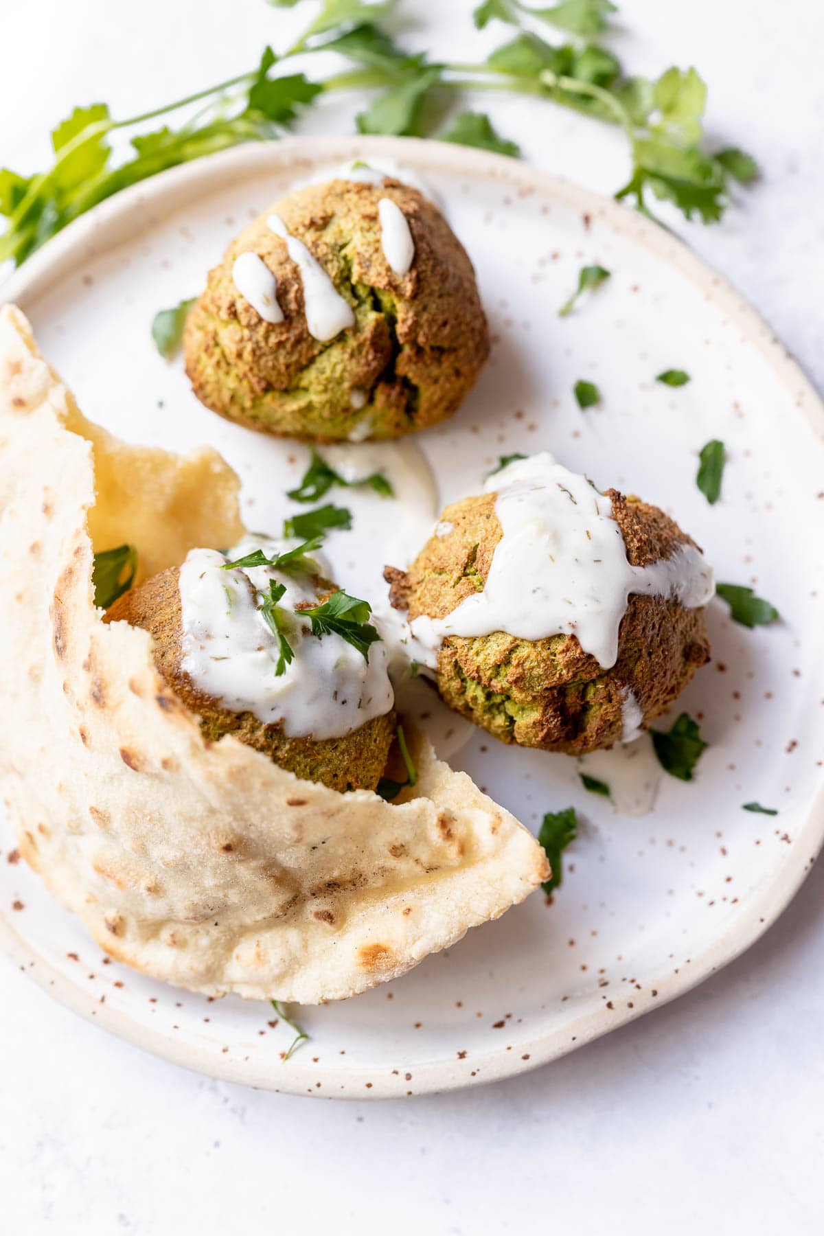 Crispy falafel balls topped with white sauce and green herbs resting on a plate with pita bread.