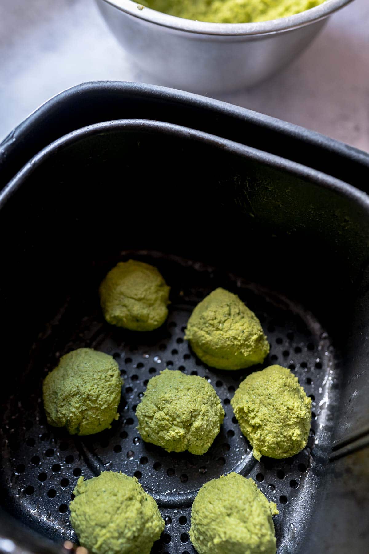 A greased air fryer basket filled with uncooked green falafel balls.
