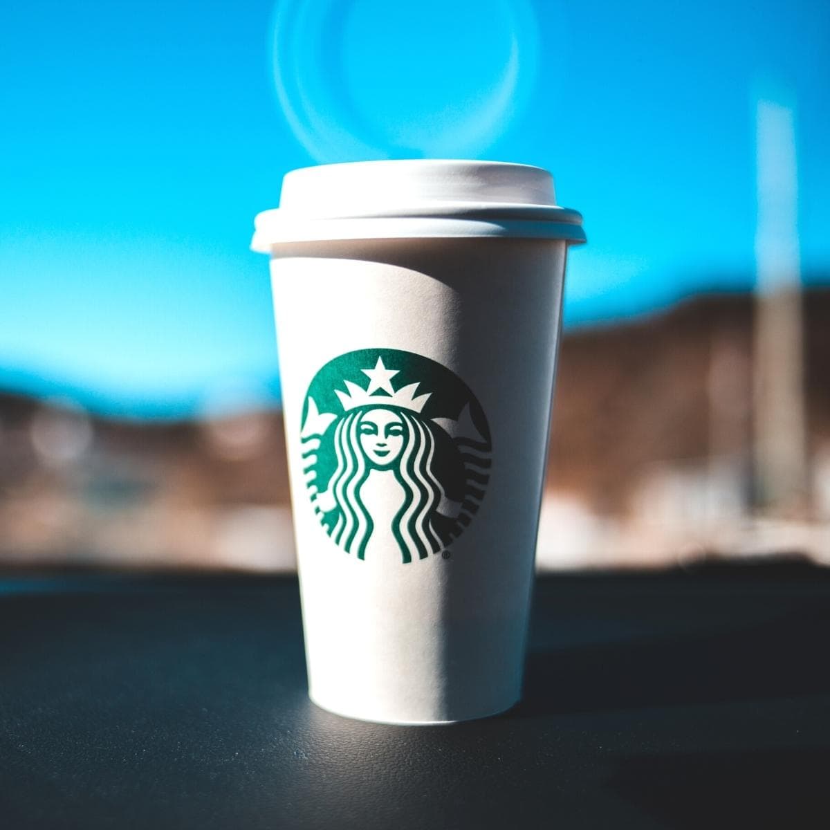 A white and green Starbucks cup resting on the dashboard of a car with a blue sky behind it.