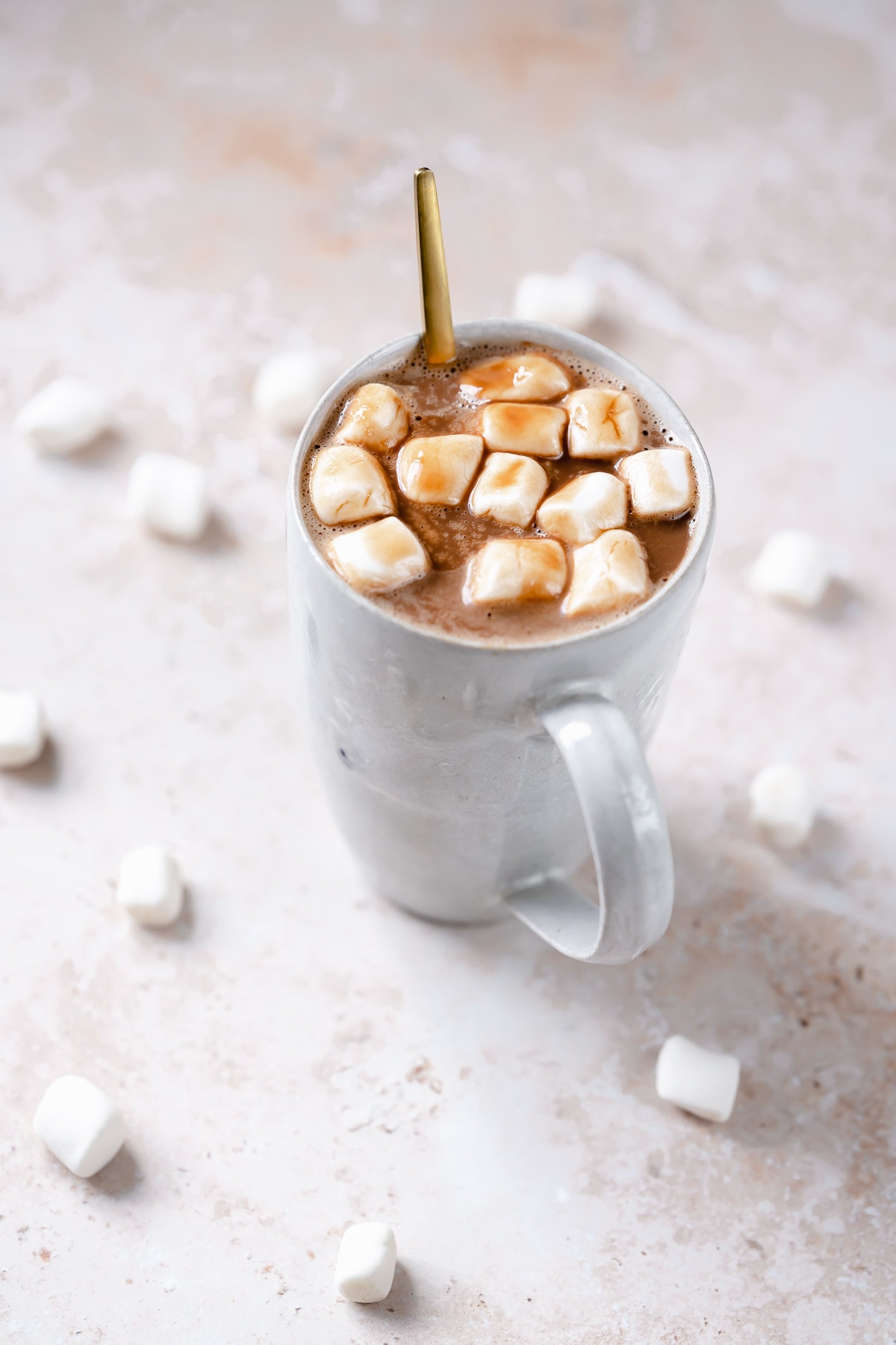 A gold spoon sticks out of a white mug filled with hot chocolate and mini marshmallows.