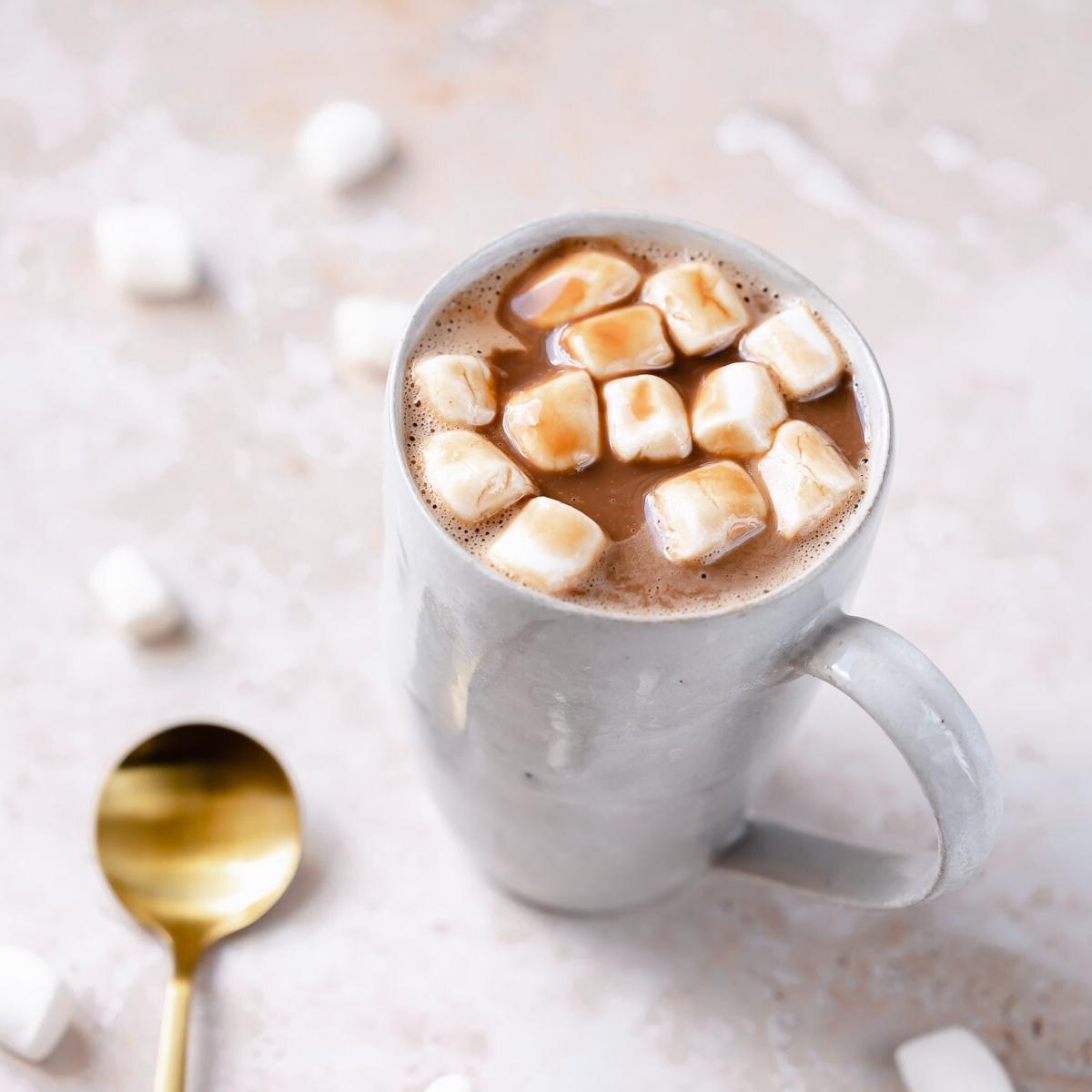 A white ceramic mug filled with brown hot chocolate and mini marshmallows resting on a tan table next to a gold spoon.