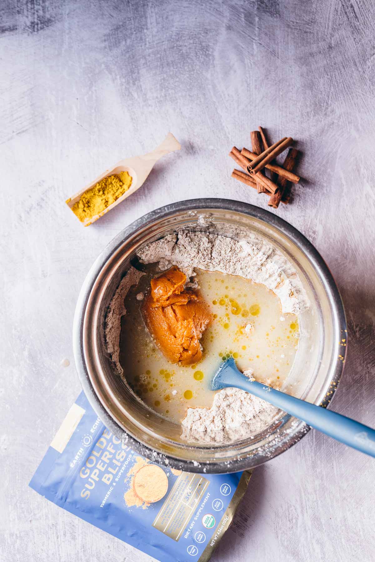 A silver mixing bowl filled with wet and dry ingredients with a blue spatula sticking out.