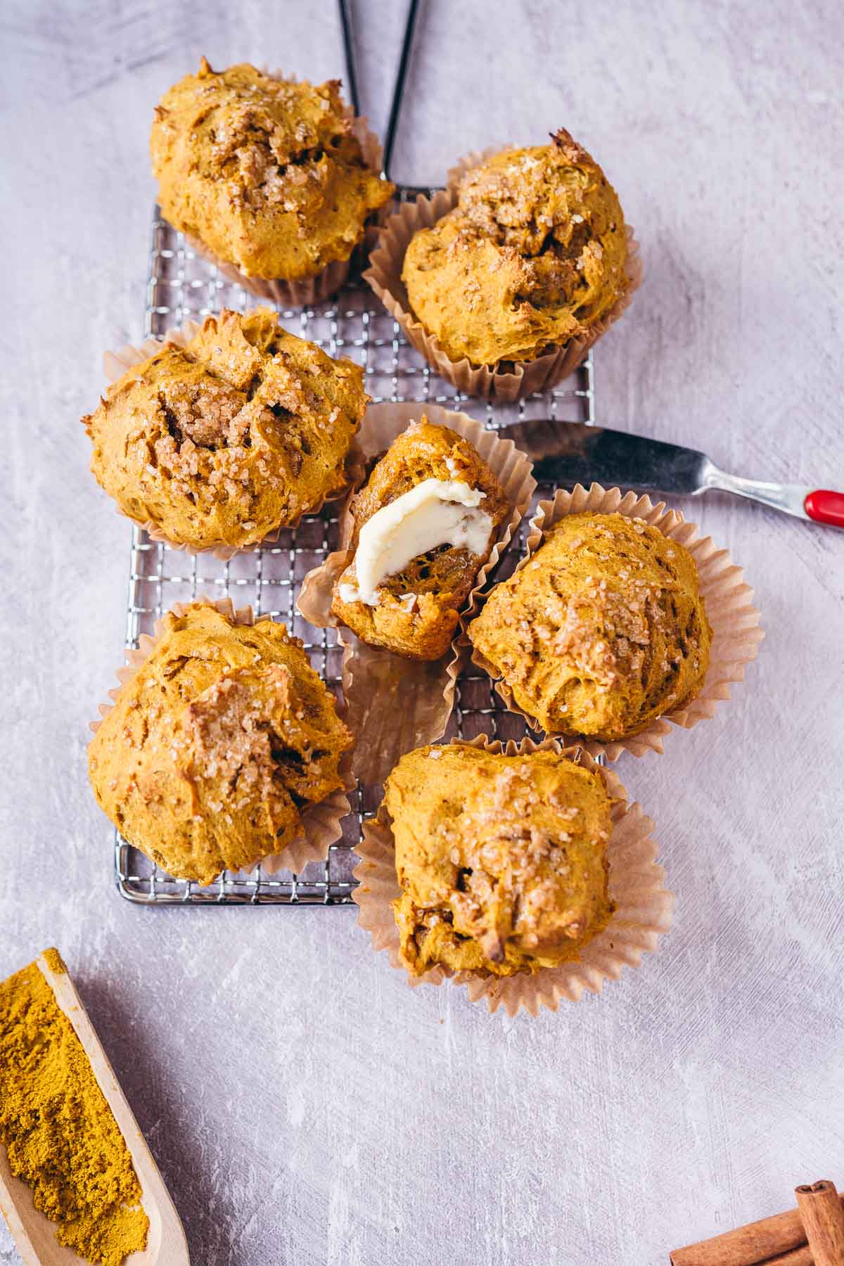 Golden muffins on a silver baking rack, one bitten into and smeared with butter by a red handled butter knife.