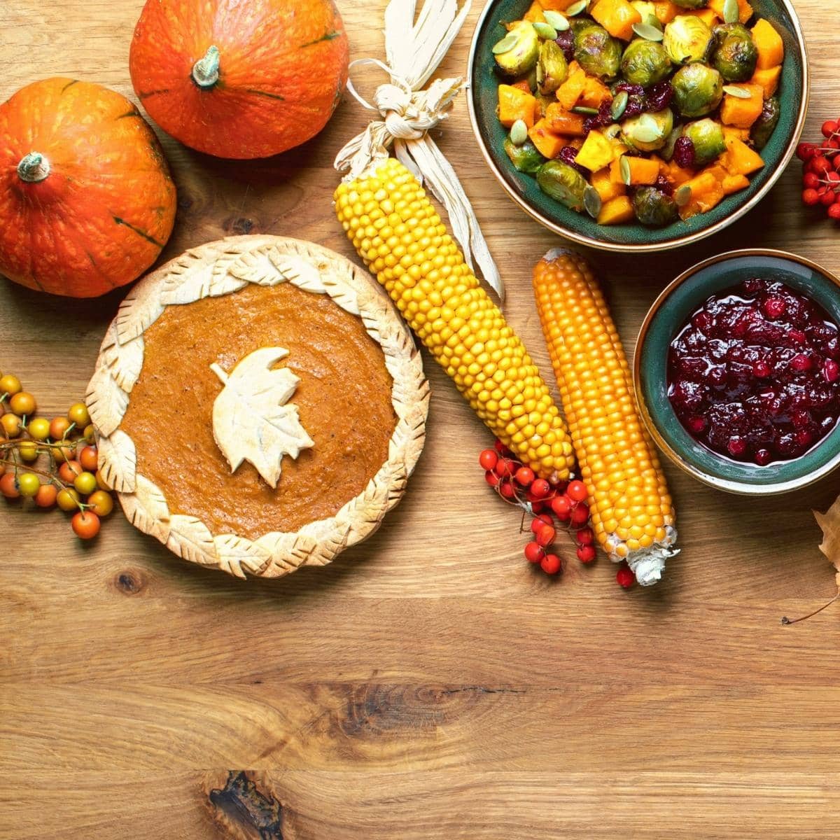 A wooden table topped with pumpkin pie, corn on the cob, cranberry sauce and butternut squash salad.