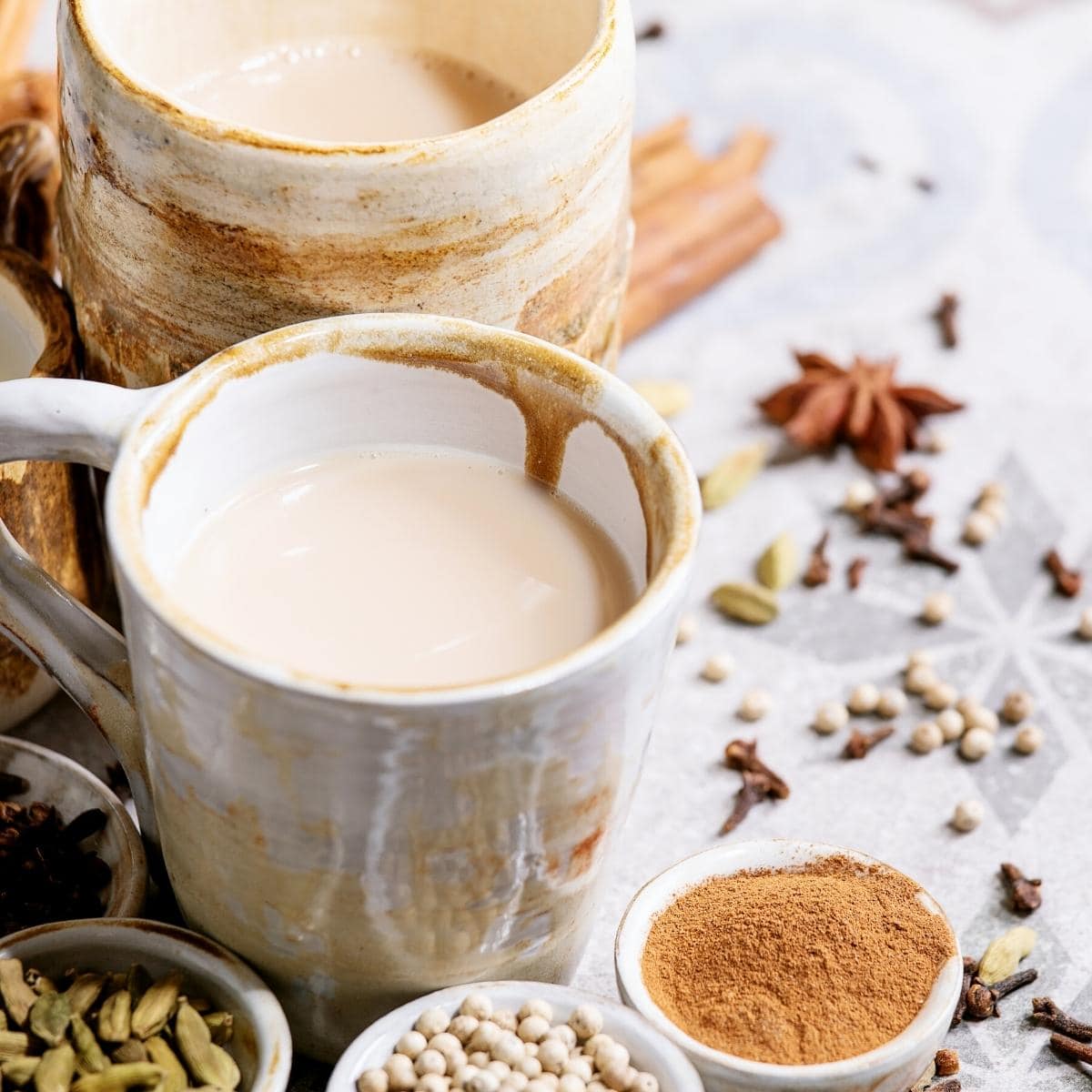 A white mug filled with chai and surround by small bowls of spices.