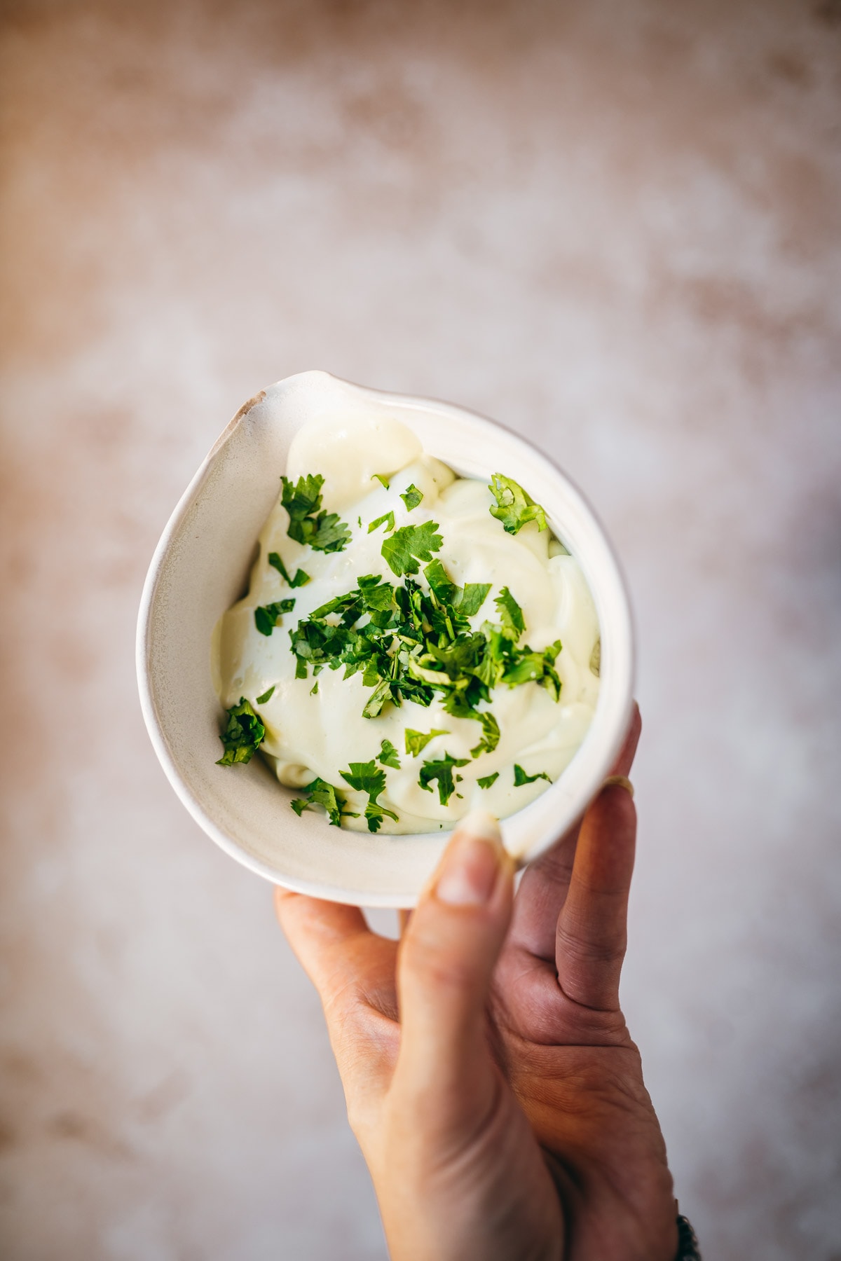 A hand holding a white ceramic bowl of creamy avocado sauce topped with fresh cilantro leaves.