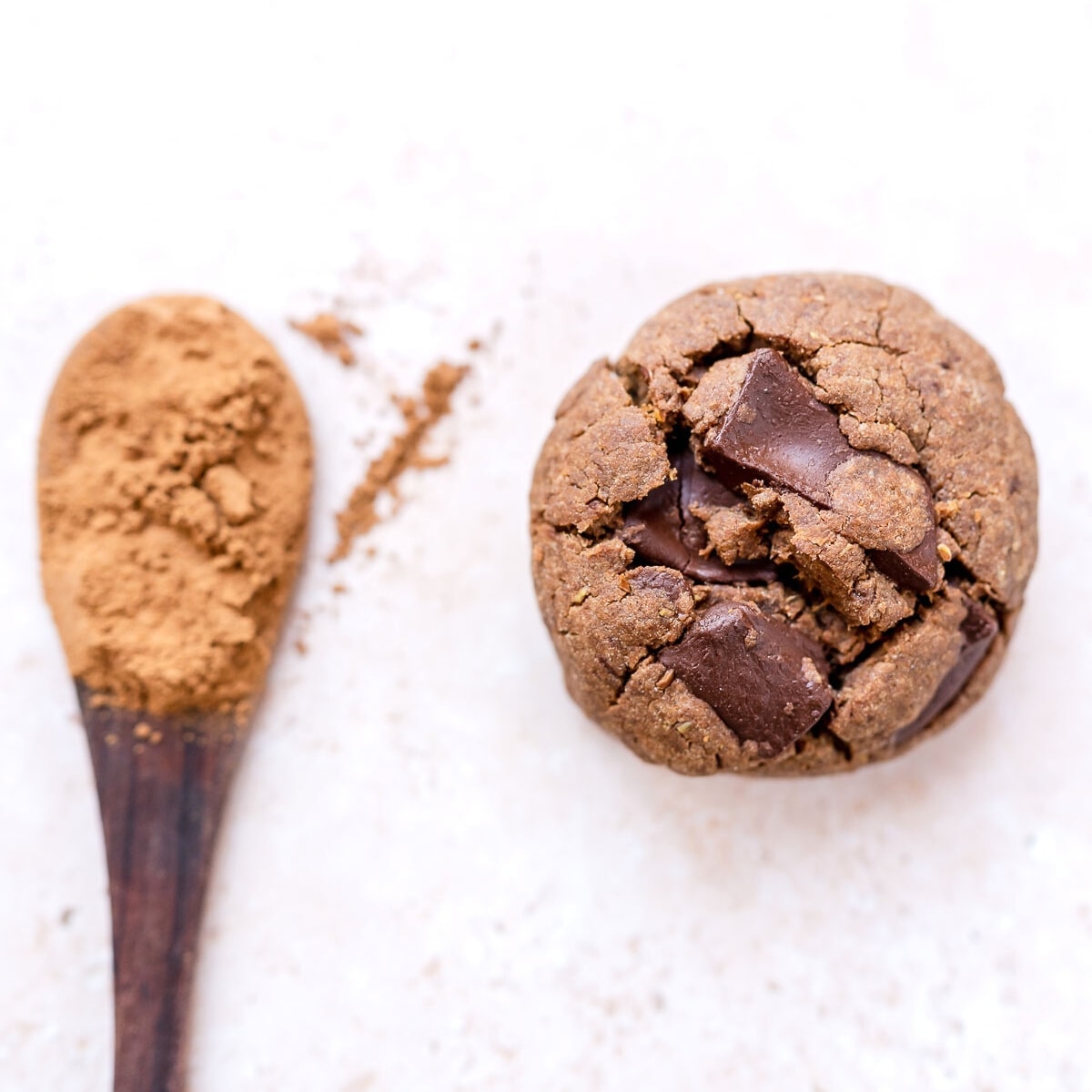 A wooden spoon filled with cacao powder rests next to a large brown chocolate cookie.