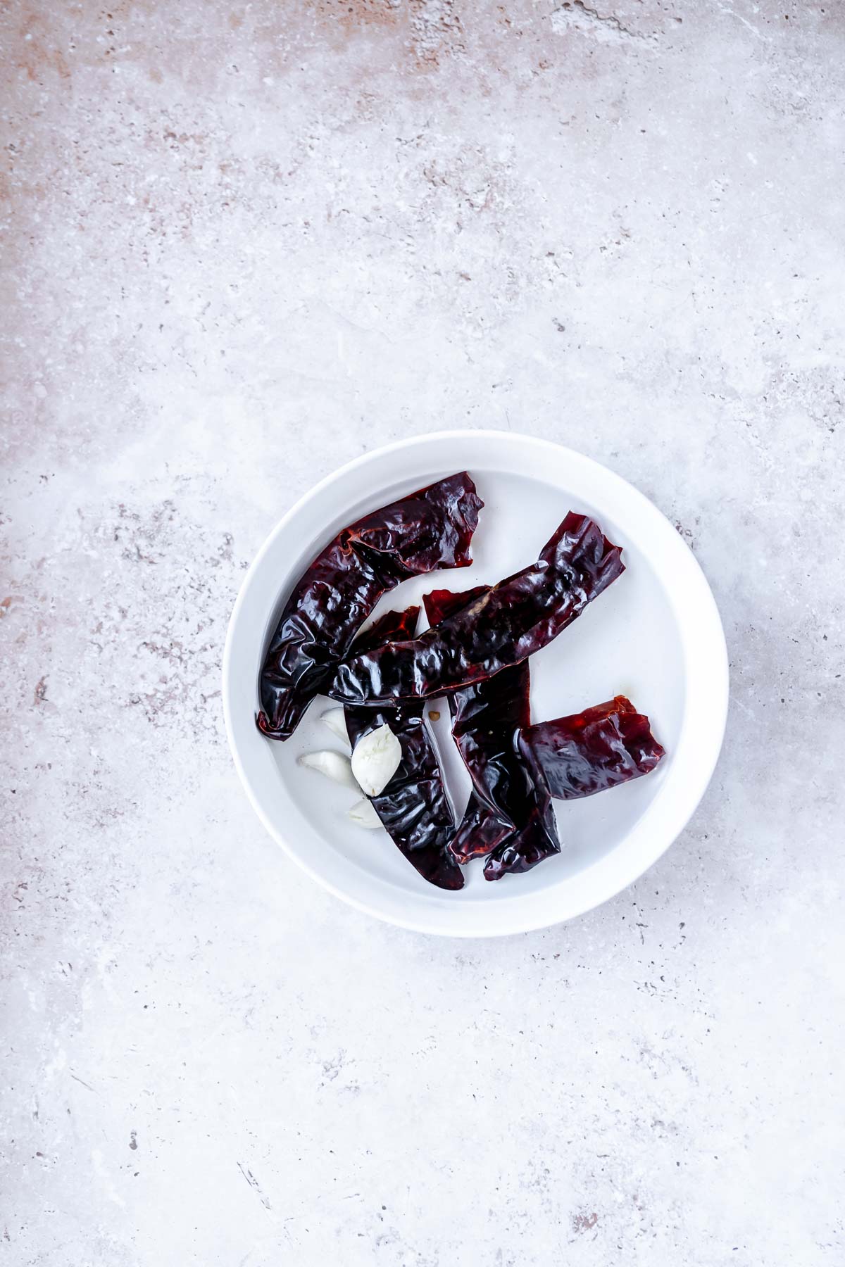 A white bowl filled with dried dark red peppers and garlic cloves.