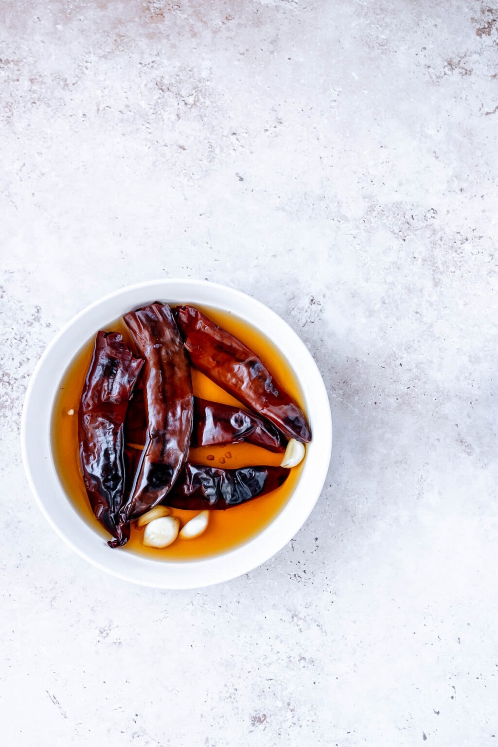 A white bowl containing dried red chilies and garlic cloves soaking in a yellowish liquid, placed on a light textured surface, evokes the vibrant beginnings of a rich Guajillo Sauce.