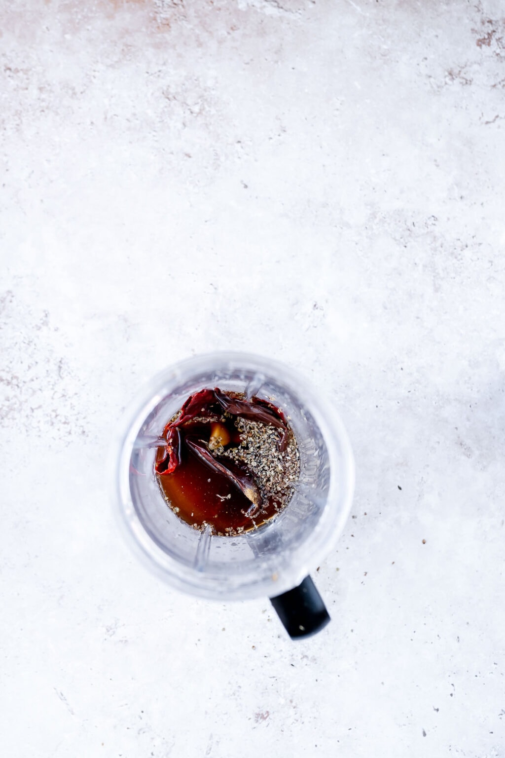 Overhead view of a blender containing Guajillo Sauce ingredients, including dried chilies, spices, and liquid, on a light textured surface.