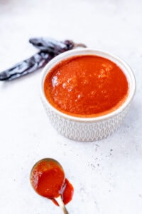 A white bowl filled with vibrant red Guajillo Sauce sits on a light surface, with a spoon of sauce and dried chili peppers nearby.