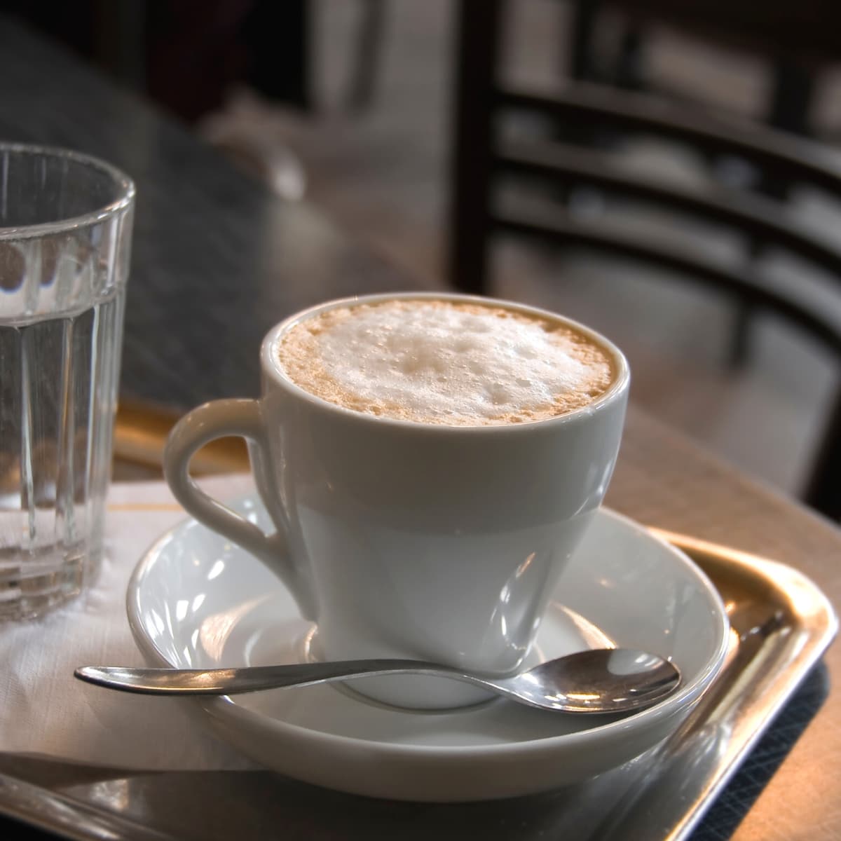 A white mug filled with Wiener Melange Austrian coffee resting on a white plate next to a silver spoon on a wood table.