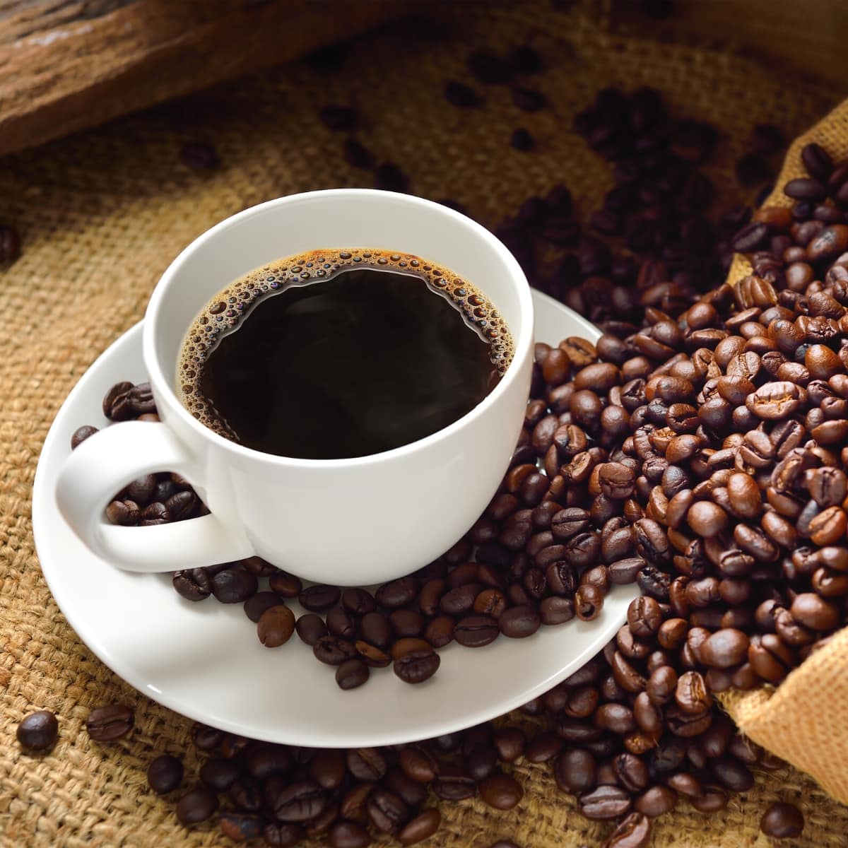 A white cup of coffee resting on a white plate surrounded by dark brown coffee beans.