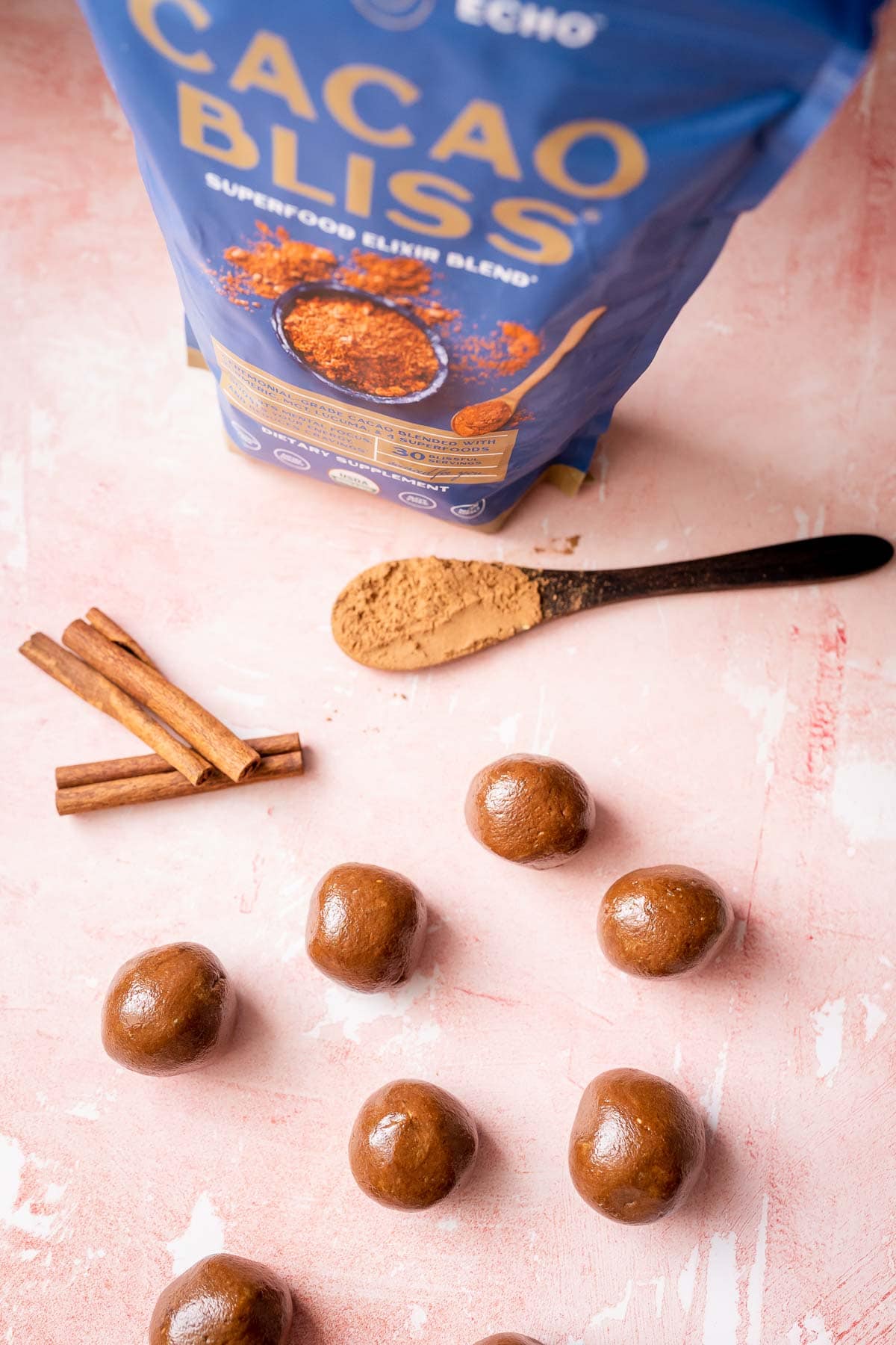A blue bag of Earth Echo Cacao Bliss rests next to brown bliss balls, cinnamon sticks and a wooden spoon.