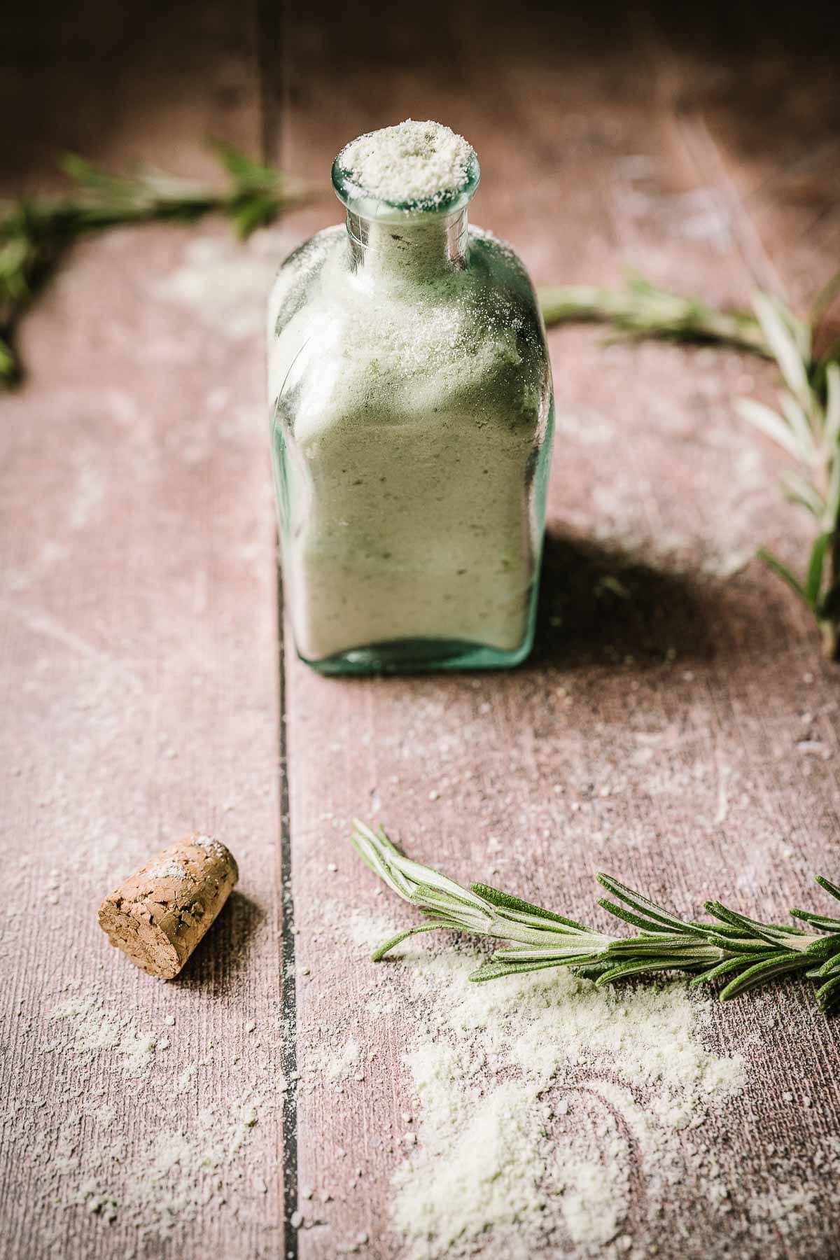 An open glass bottle filled with light green salt resting on a light wood tabletop.