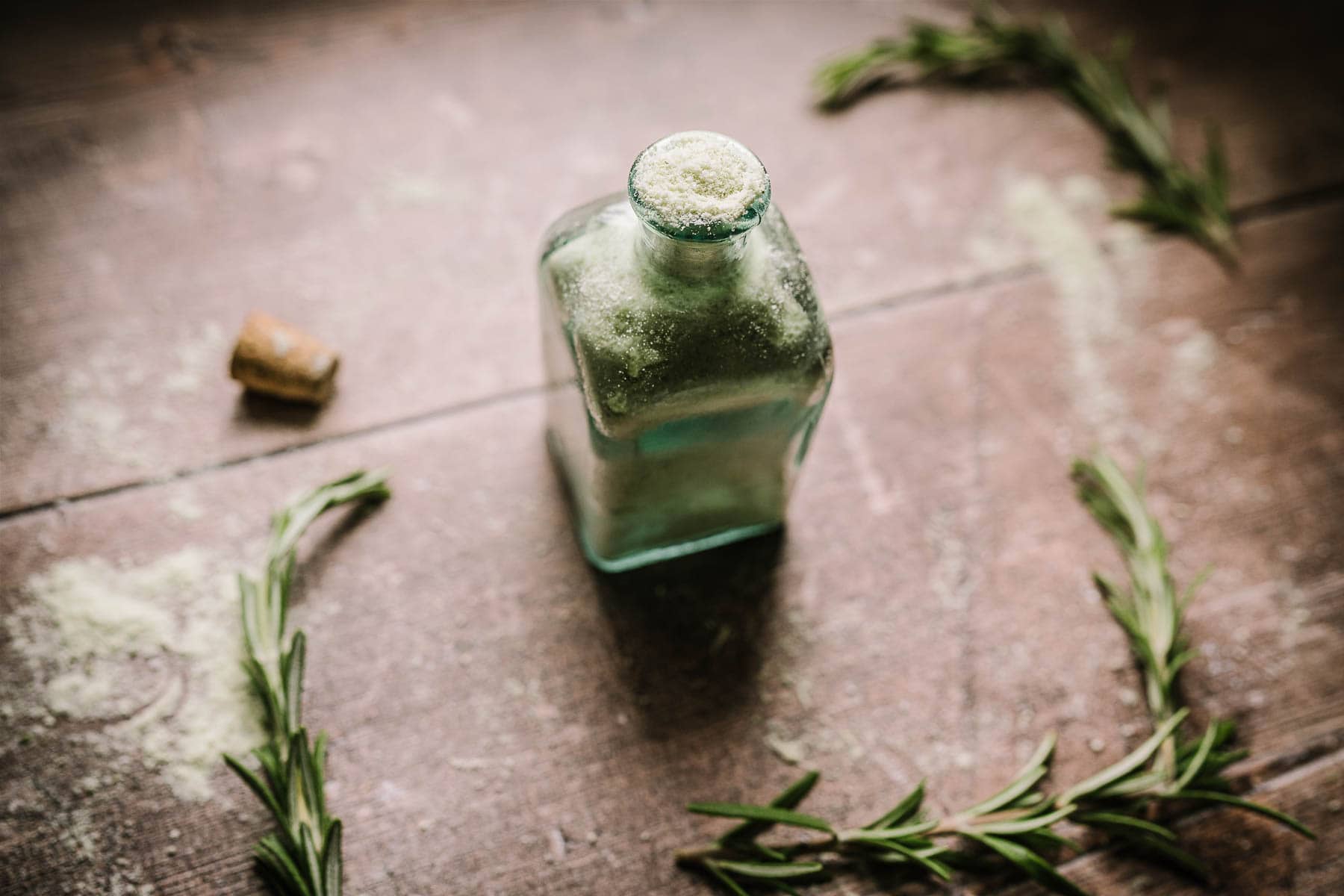 An open glass bottle filled with light green salt resting on a wooden table scattered with fresh rosemary sprigs.