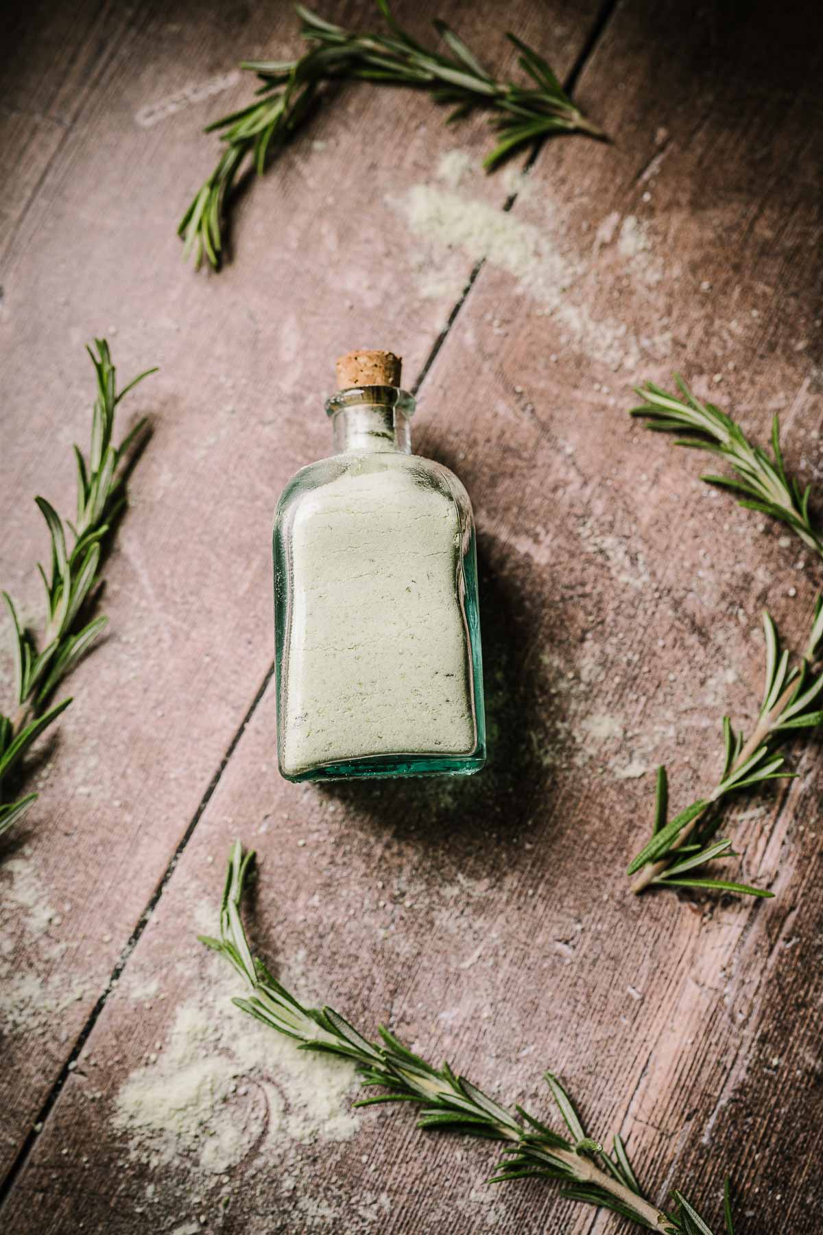 A corked glass bottle filled with light green salt resting on a wooden table scattered with rosemary sprigs.