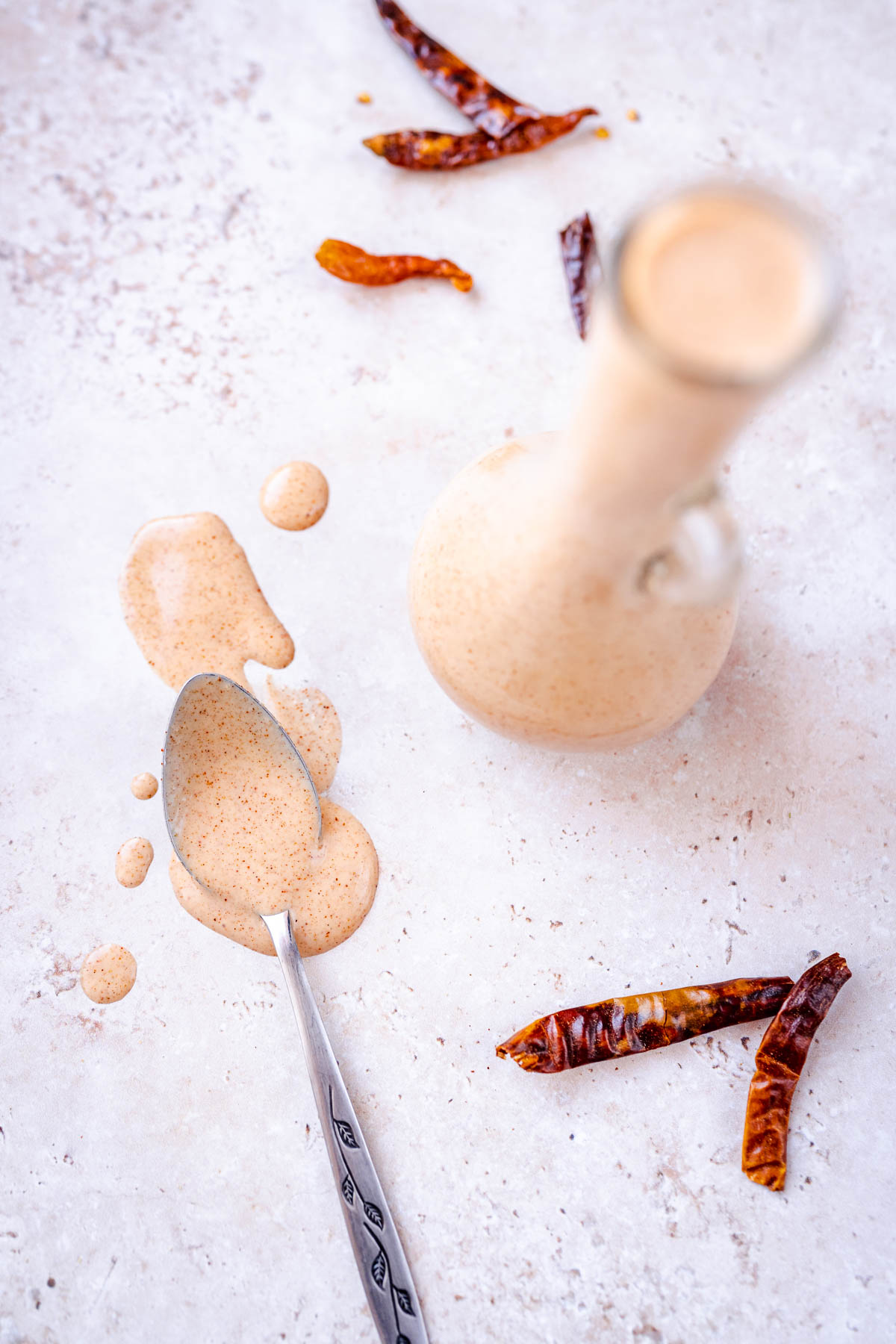 A silver spoon dripping with homemade salad dressing resting next to a bottle of dressing and dried red chiles.