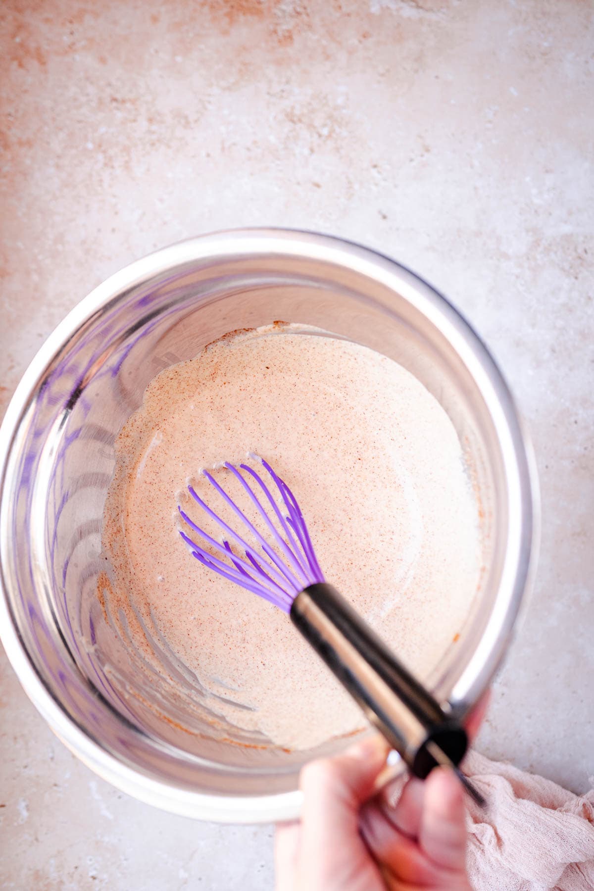 A purple whisk stuck in a light red sauce in a silver mixing bowl.
