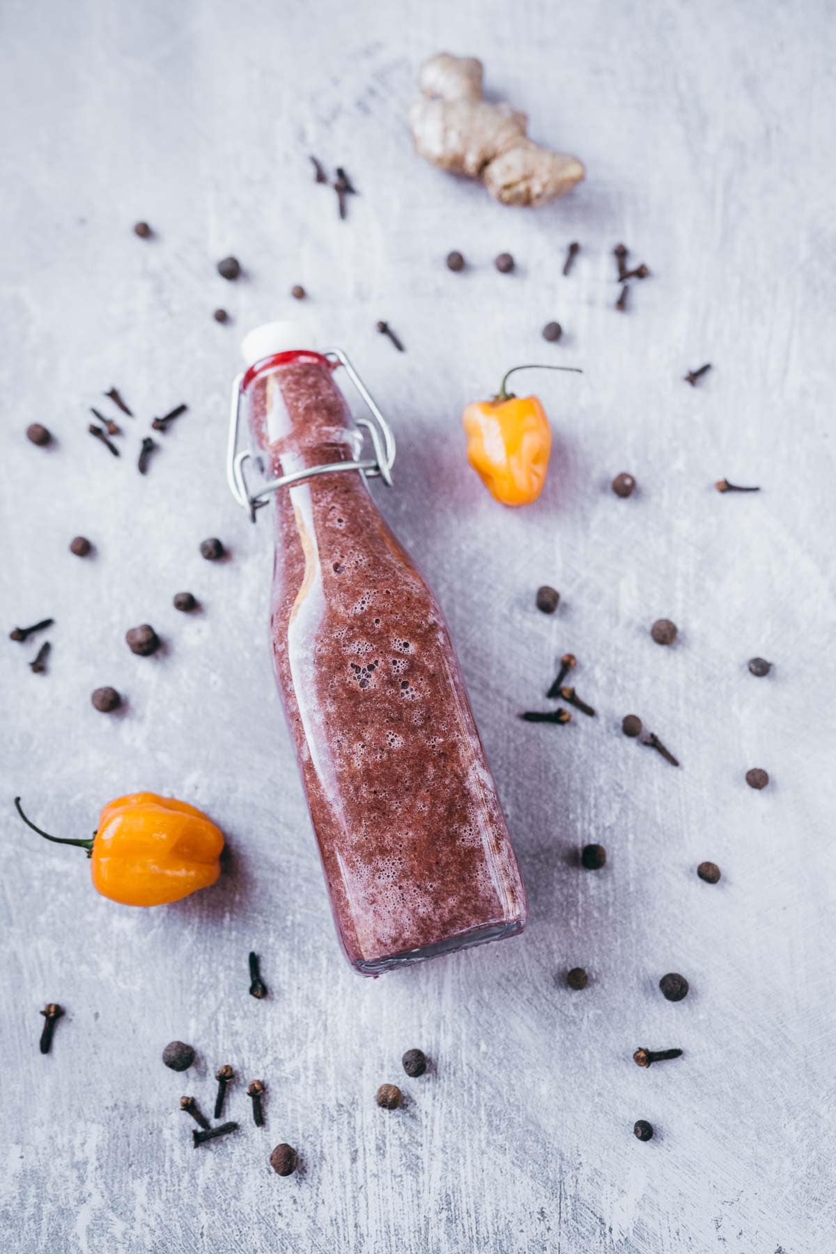 A bottle of hot sauce resting on a gray table scattered with dry spices and orange chili peppers.