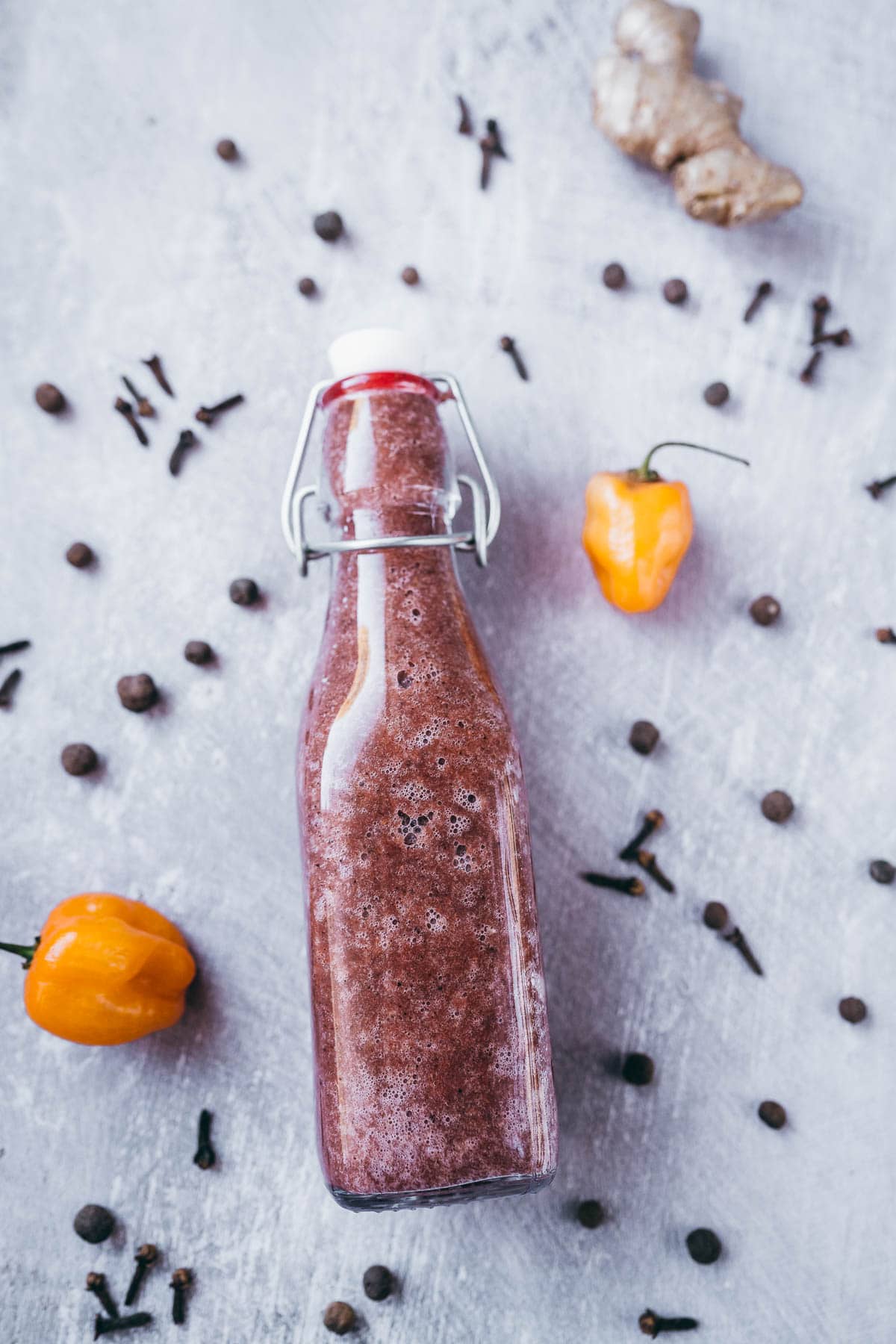 A tall glass bottle filled with a reddish brown sauce resting on a gray table scattered with peppers and spices.