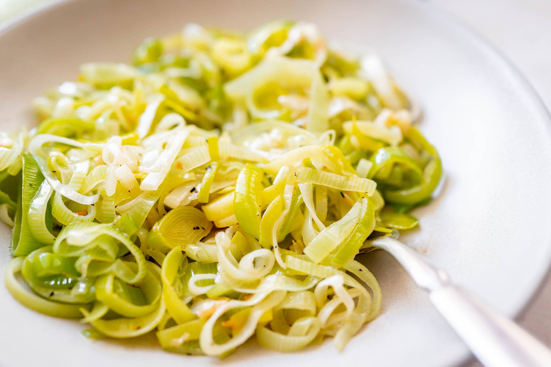 Tender, sliced, buttered leeks in a large ceramic bowl with a silver fork.