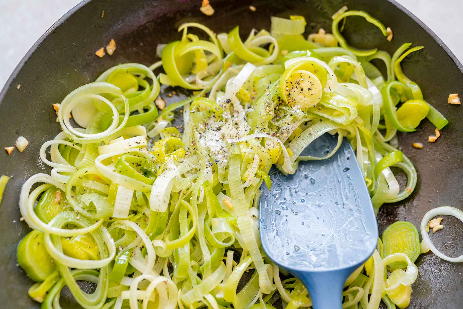 A blue silicone spatula rests in a large skillet of sliced leeks, salt and pepper.
