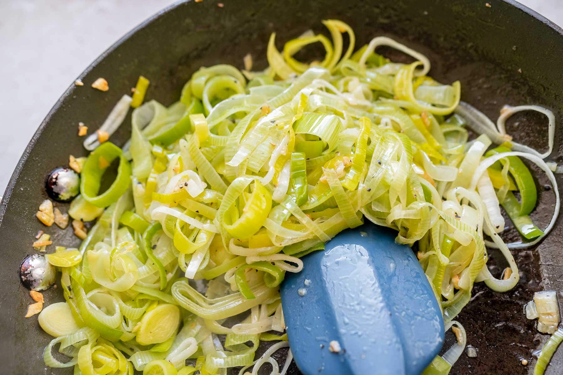 Freshly sauteed leeks rest in a dark black skillet with a blue spatula.