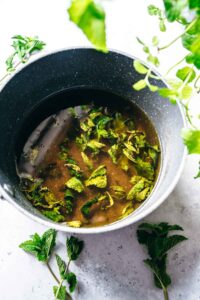 A pot filled with brown mint simple syrup and floating mint leaves, surrounded by fresh mint sprigs on a light surface.