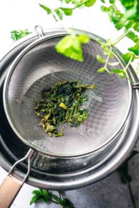 A fine mesh strainer with wet, wilted green leaves sits over a metal bowl, surrounded by fresh mint sprigs—perfect for making homemade mint simple syrup.