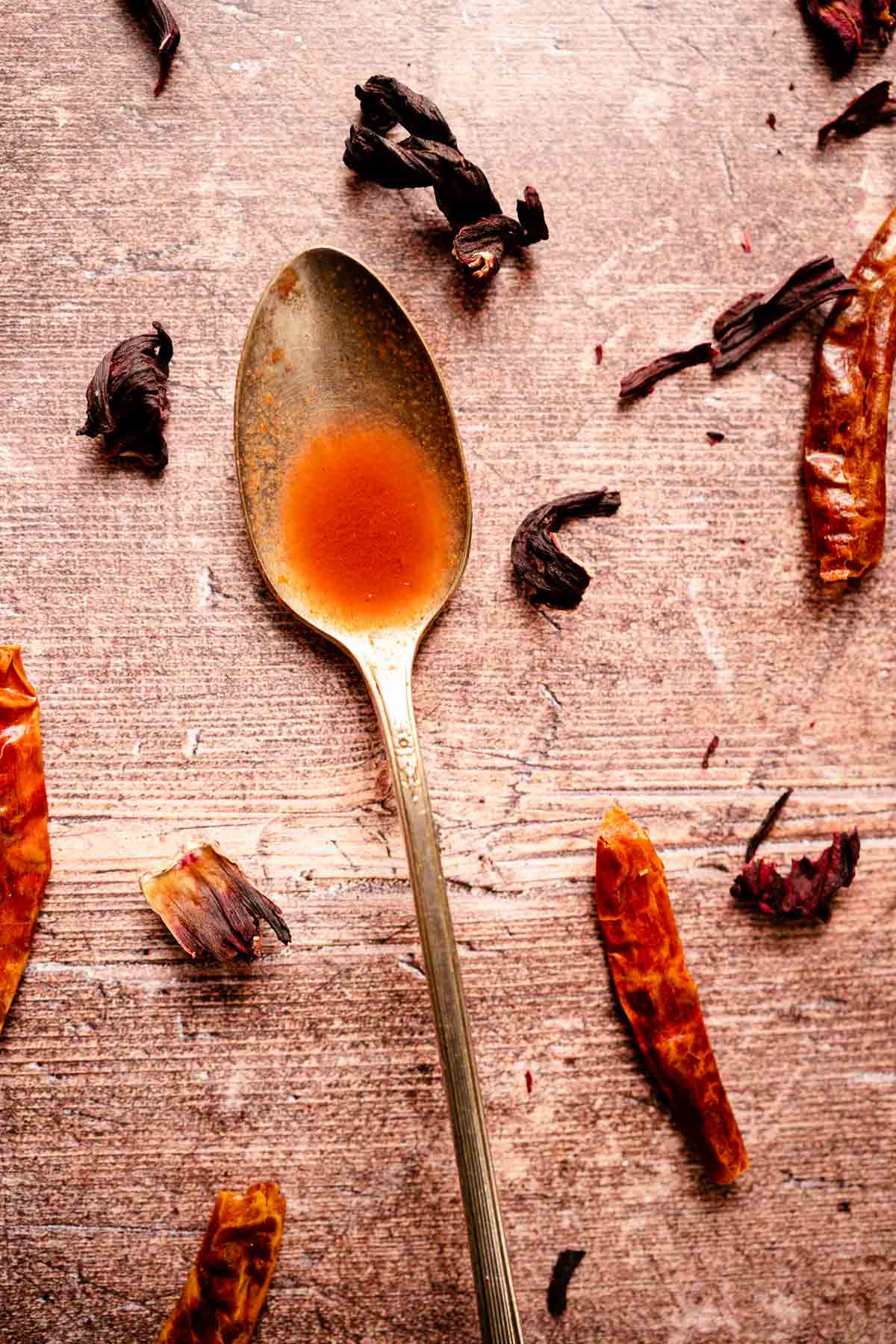 A silver spoon filled with red hot sauce resting on a wood table scattered with dried peppers and flowers.