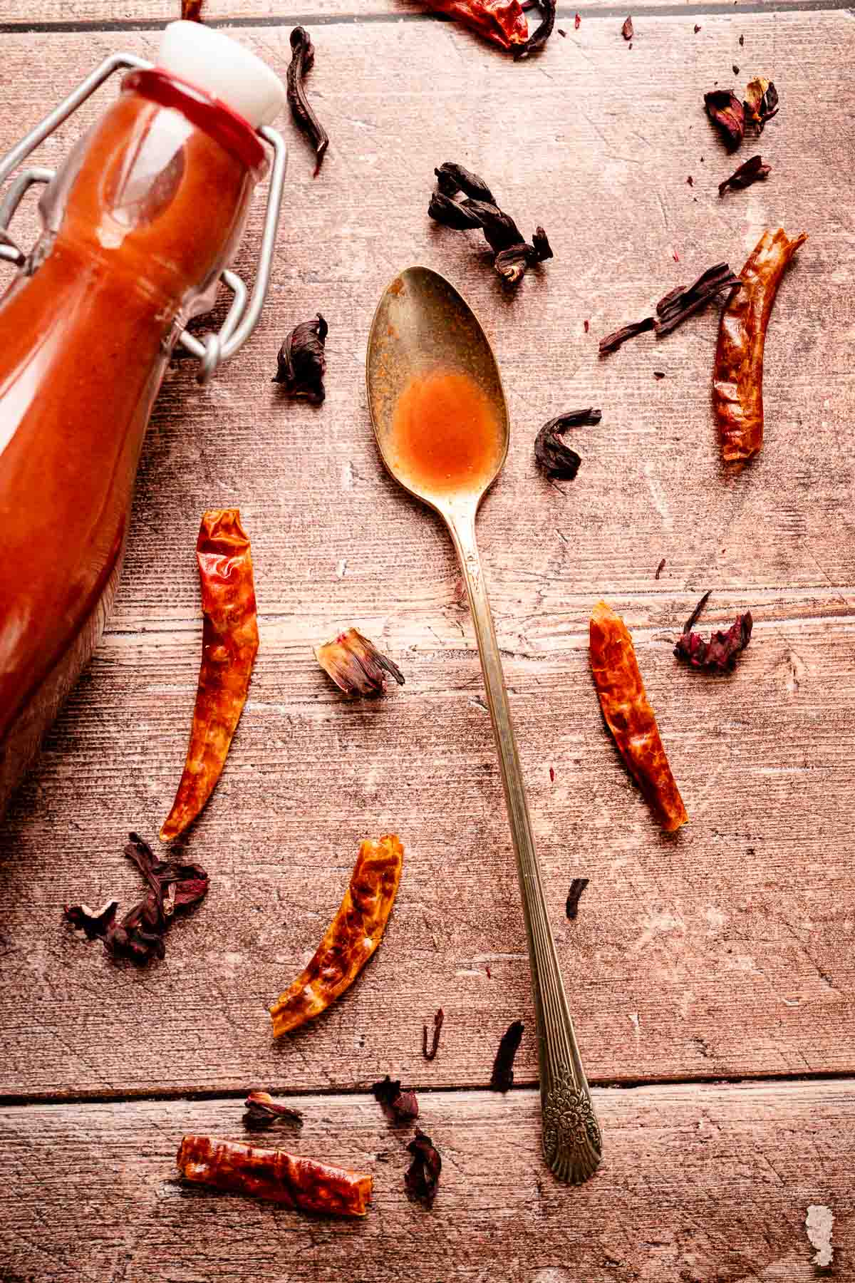 A wooden table scattered with dried chiles and hibiscus flowers, a bottle of homemade hot sauce and silver spoon holding red sauce.