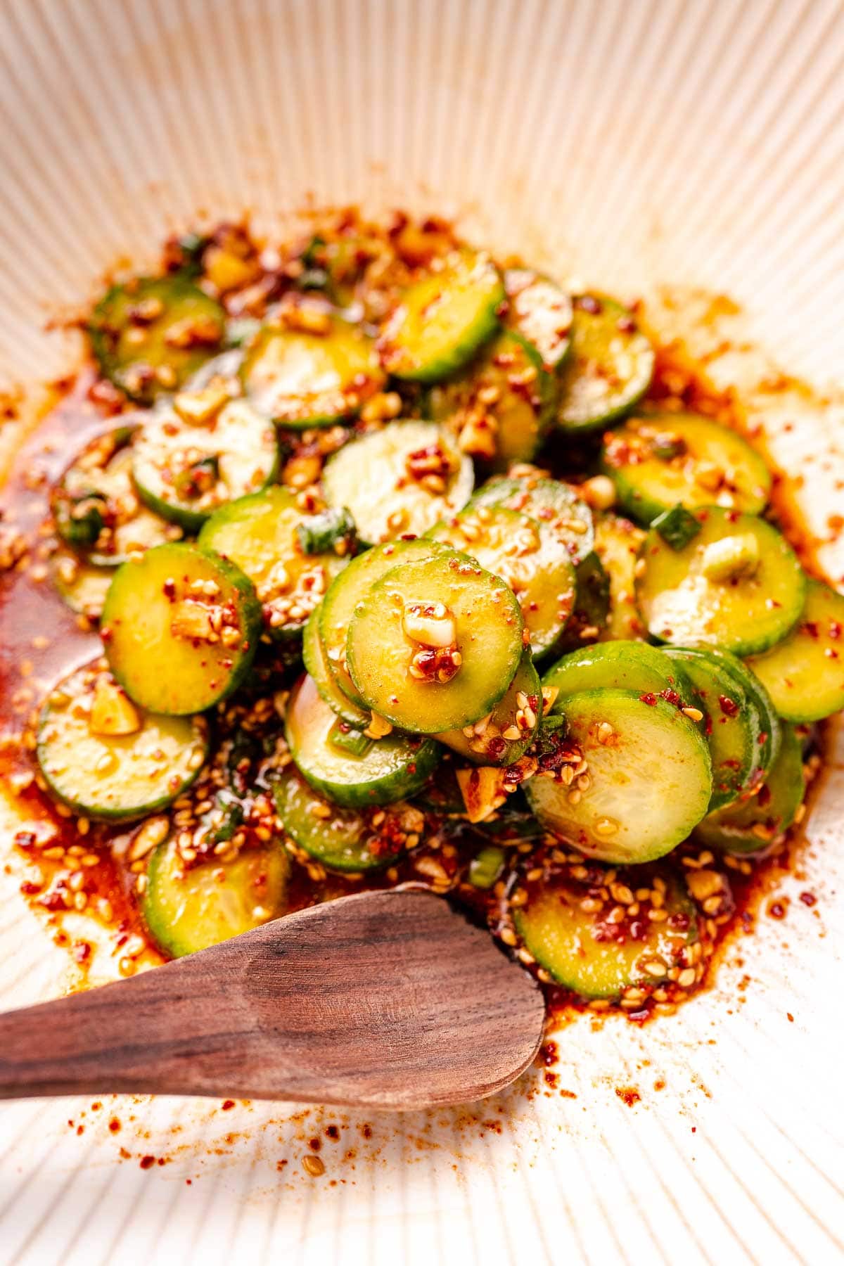 A wooden spoon rests in a white bowl filled with a cucumber salad.