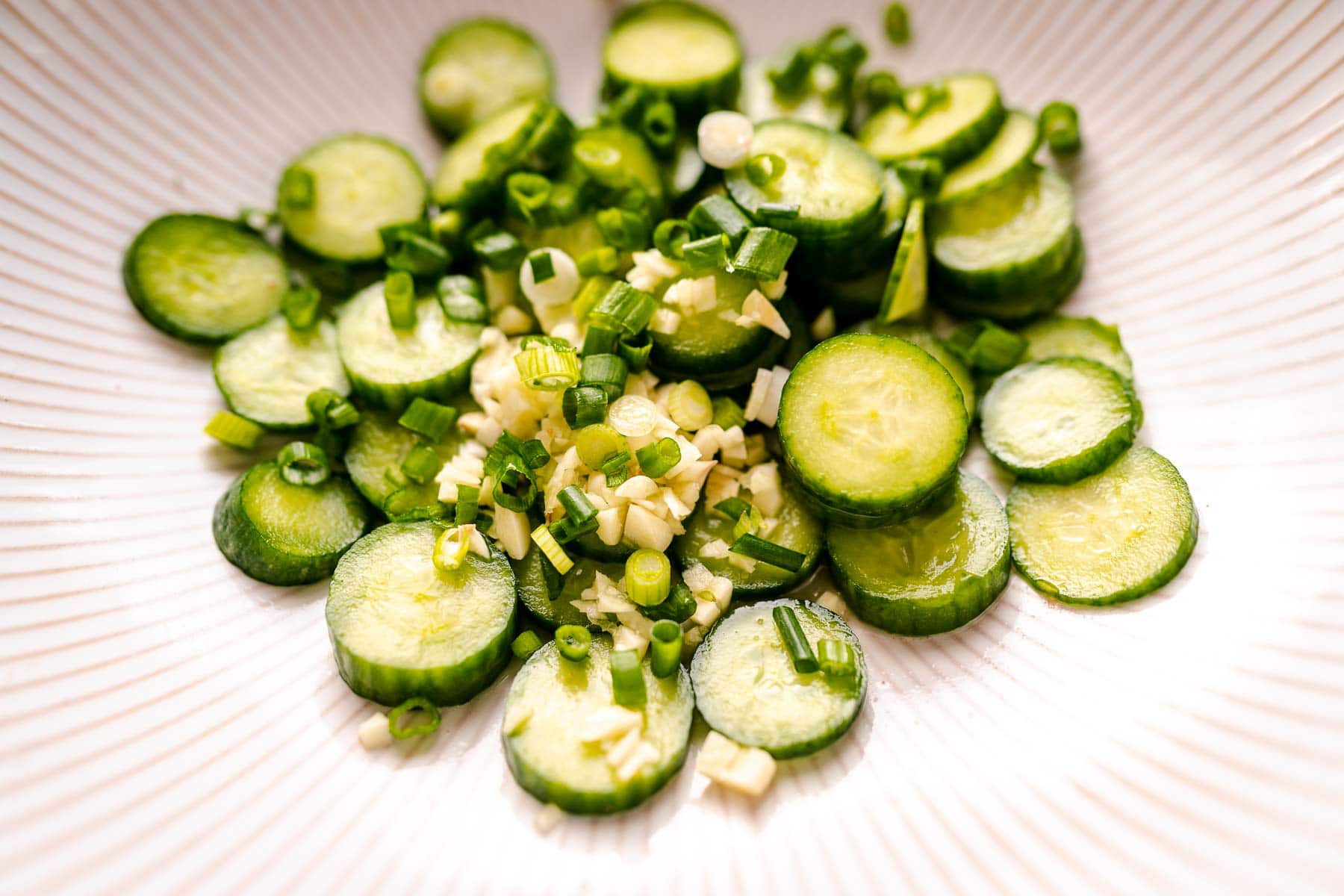 Slices of cucumber in a white bowl with sliced green onions and garlic.